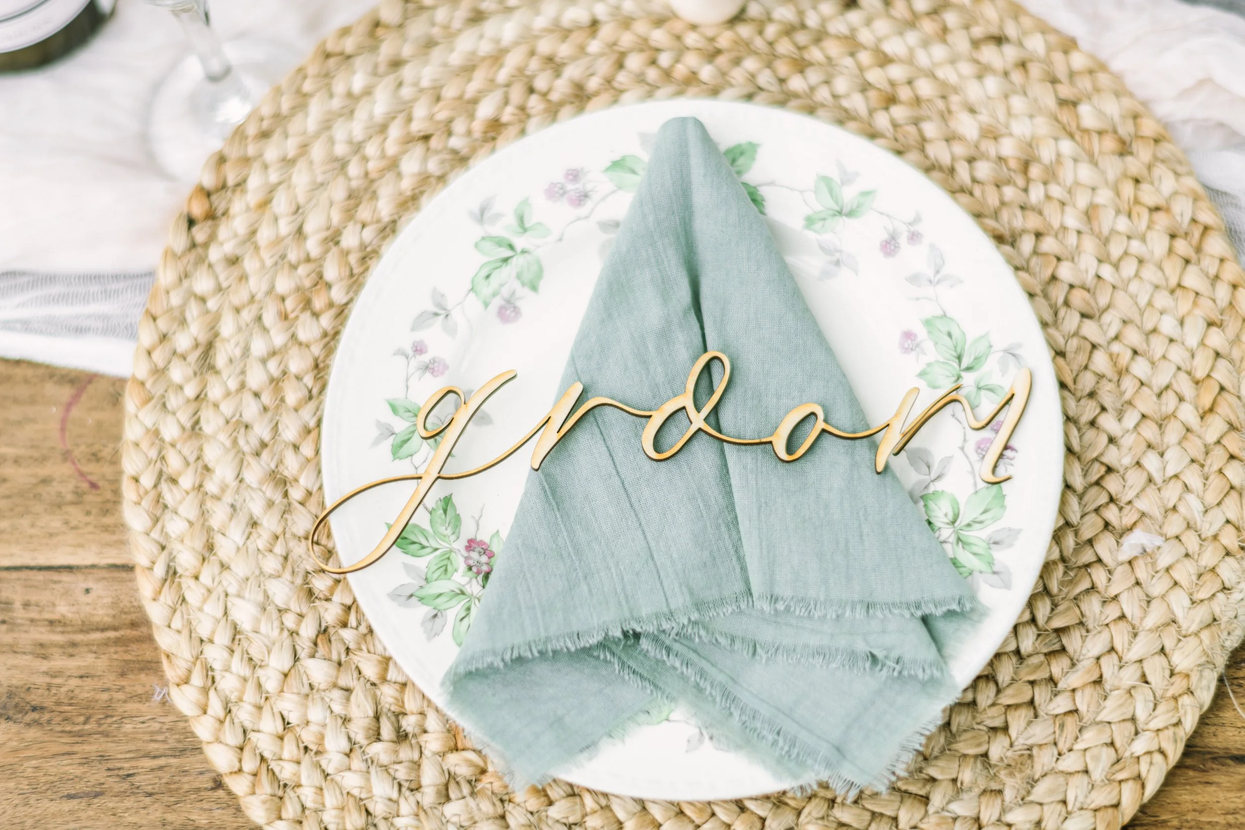 Table setting with a floral plate, green napkin, and a gold "Groom" sign on a woven table mat.
