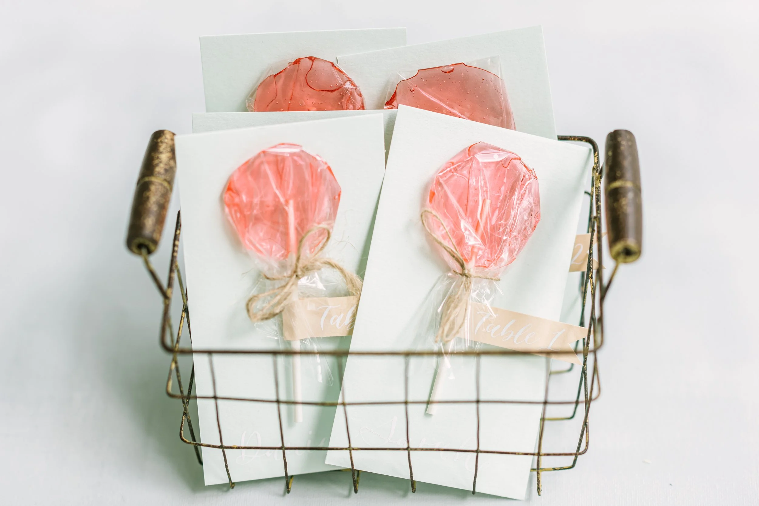 A small wire basket contains four pink lollipops wrapped in clear plastic, each secured with a beige twine bow, and individual white cards underneath.