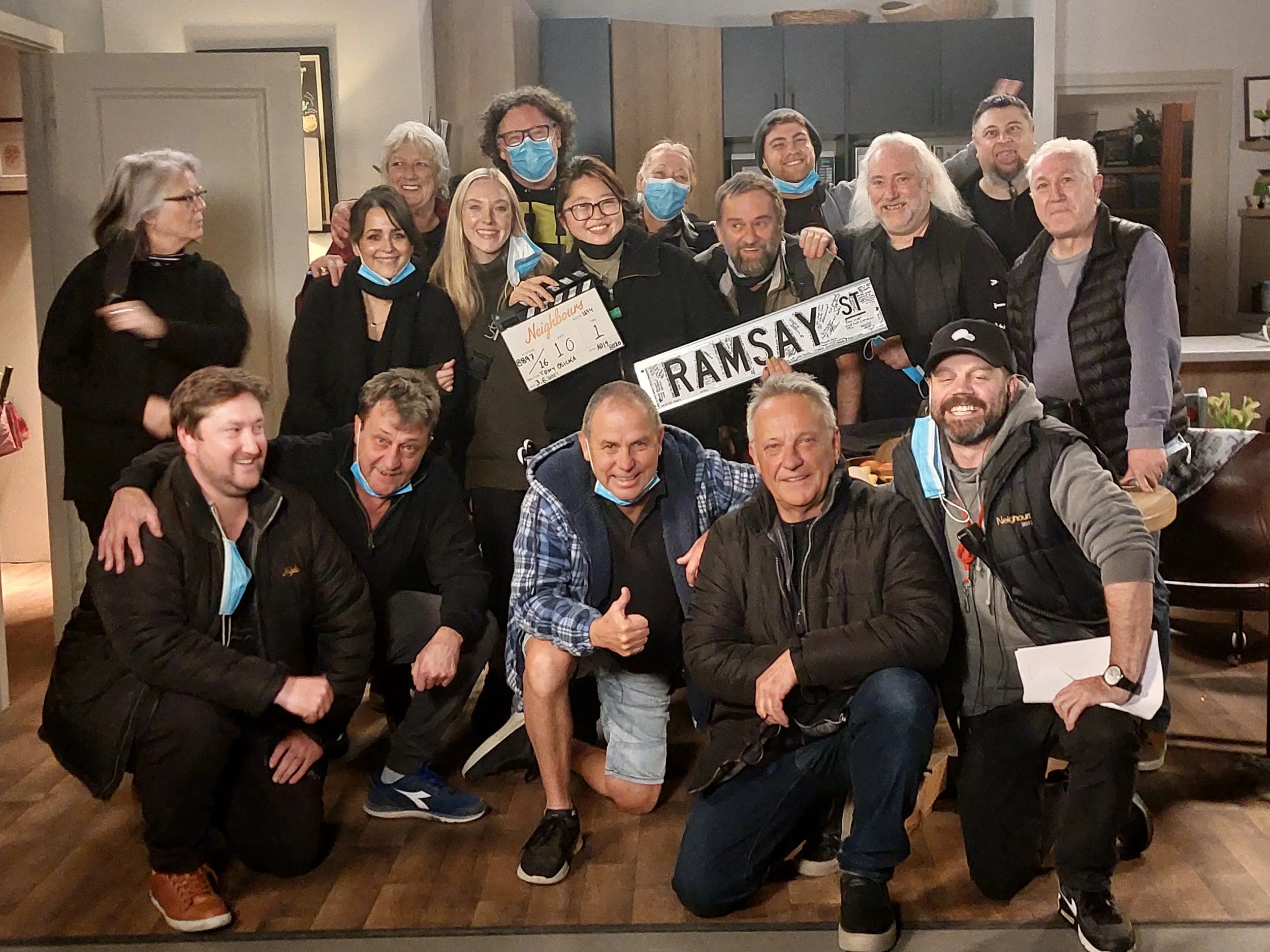 A group of people, some wearing face masks, pose together inside a kitchen for a photo. They are holding a sign that says 'Ramsay Street' and a clapperboard with 'Neighbours' written on it. Everyone appears happy and excited.