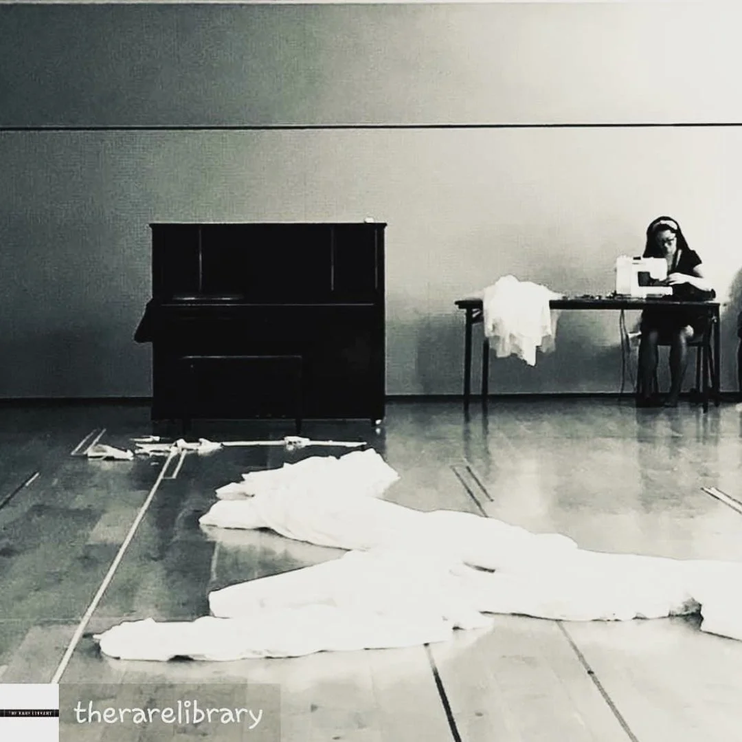An empty room with a closed piano on the left, a empty table with white fabric on it, a woman working on a sewing machine on the right, and broken white fabric or paper on the floor.