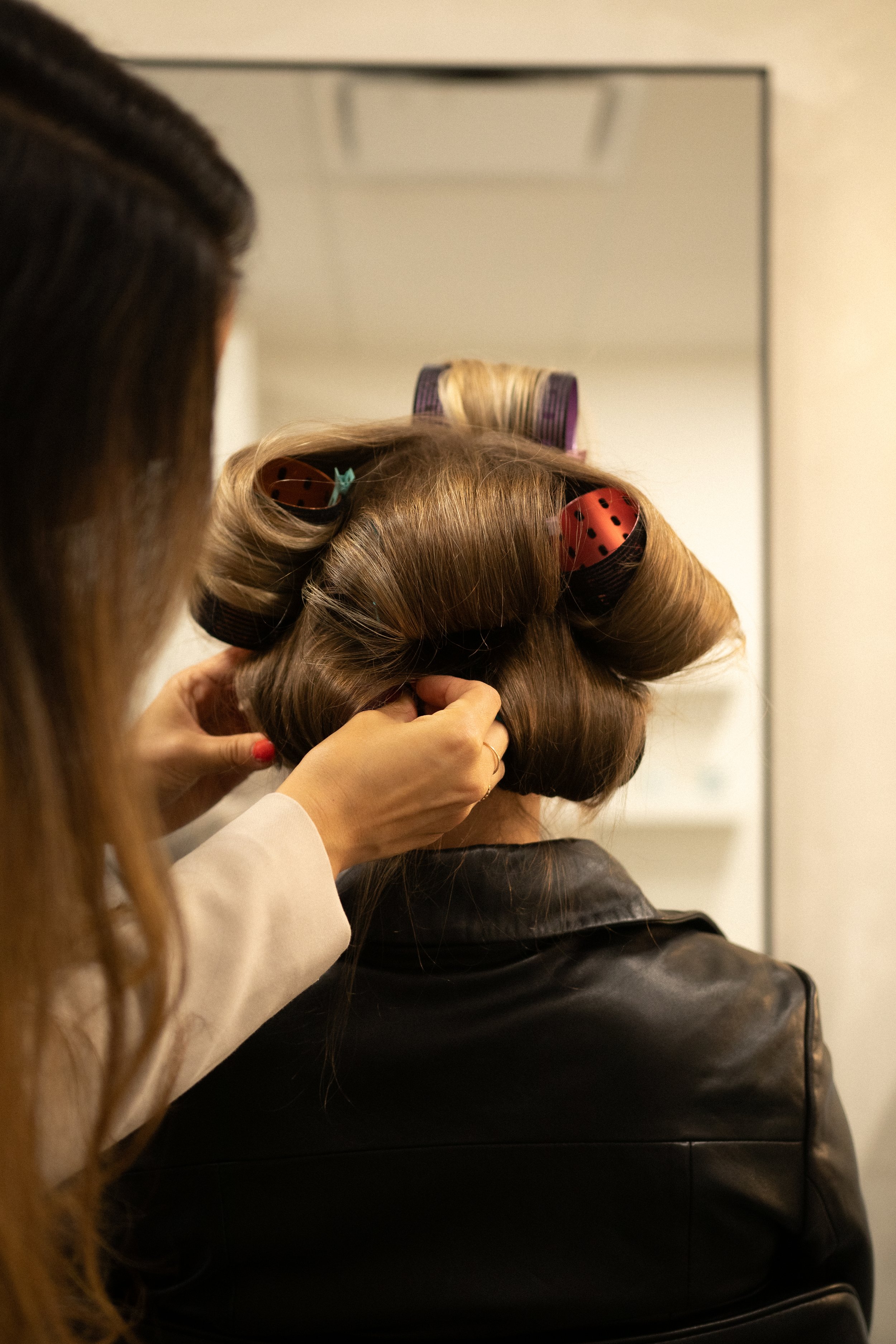 A person with light brown hair using large red and black hair rollers, assisted by another person, in front of a mirror.