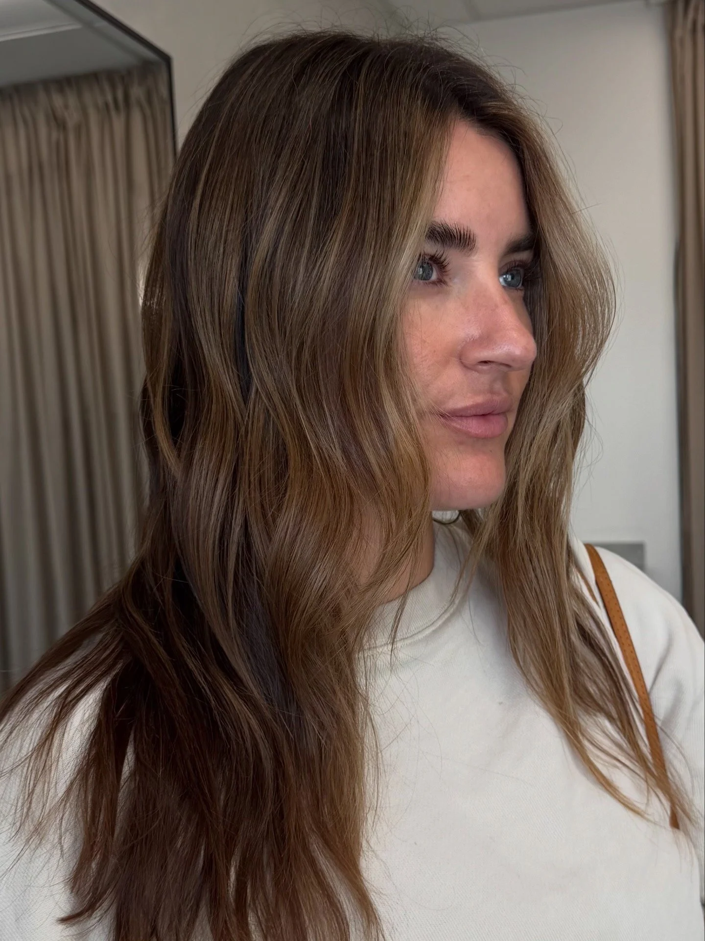 Close-up of a woman with long, wavy brown hair and blue eyes, wearing a white top, in a room with beige curtains and a mirror in the background.