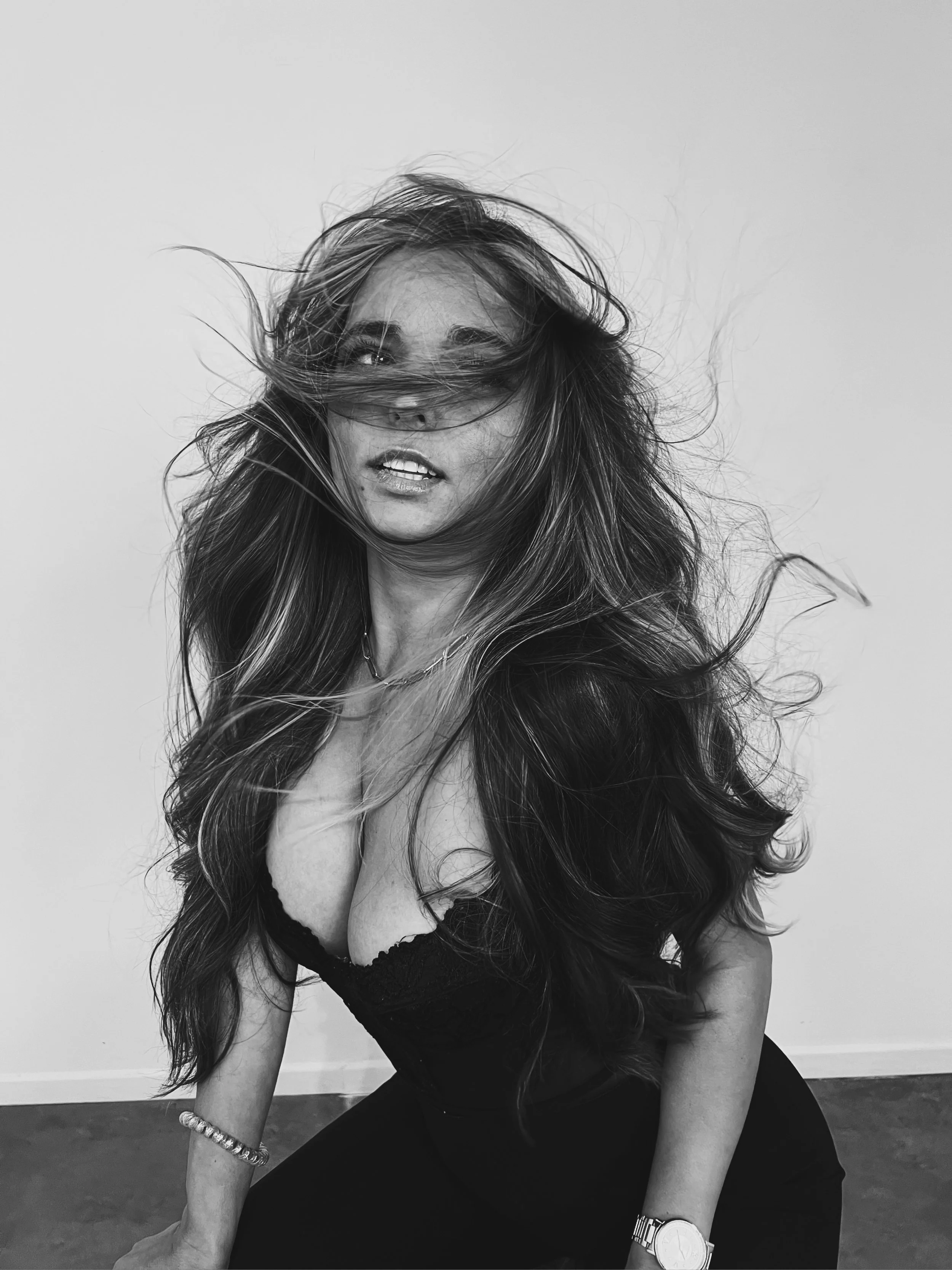 Black and white photo of a woman with long hair covering part of her face, wearing a black lace top, bracelets and a watch, sitting on the floor against a plain wall.