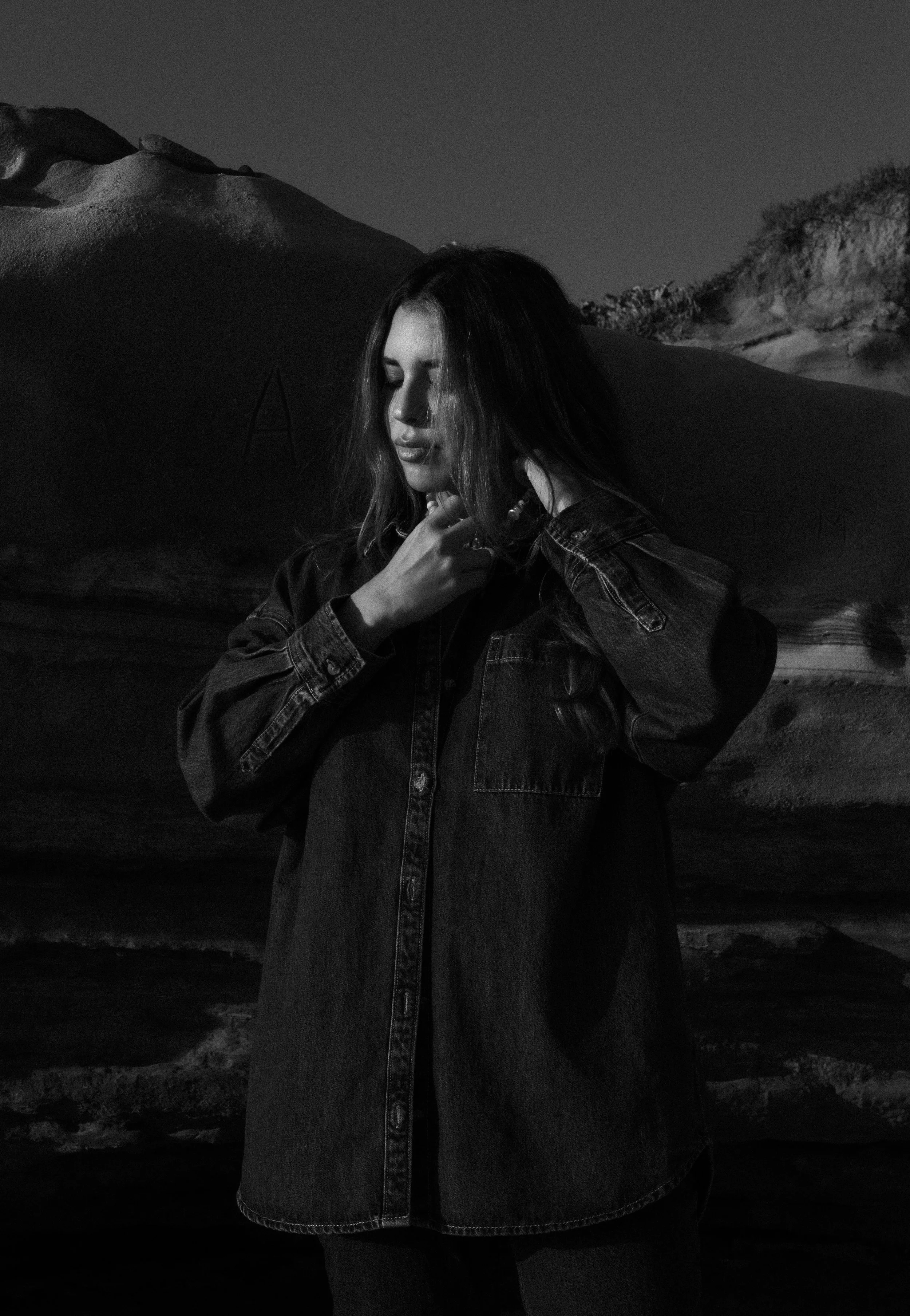 Black and white photo of a woman with long hair, wearing a denim jacket, standing outdoors at night with sand dunes in the background.
