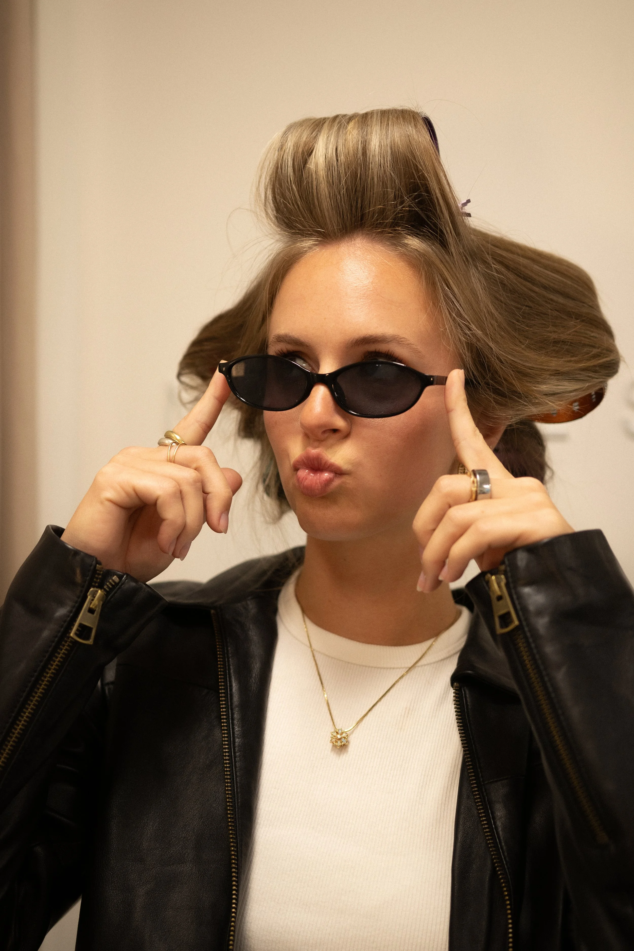 Young woman with blonde hair wearing black sunglasses, a white top, gold jewelry, and a black leather jacket, adjusting her sunglasses with both hands.