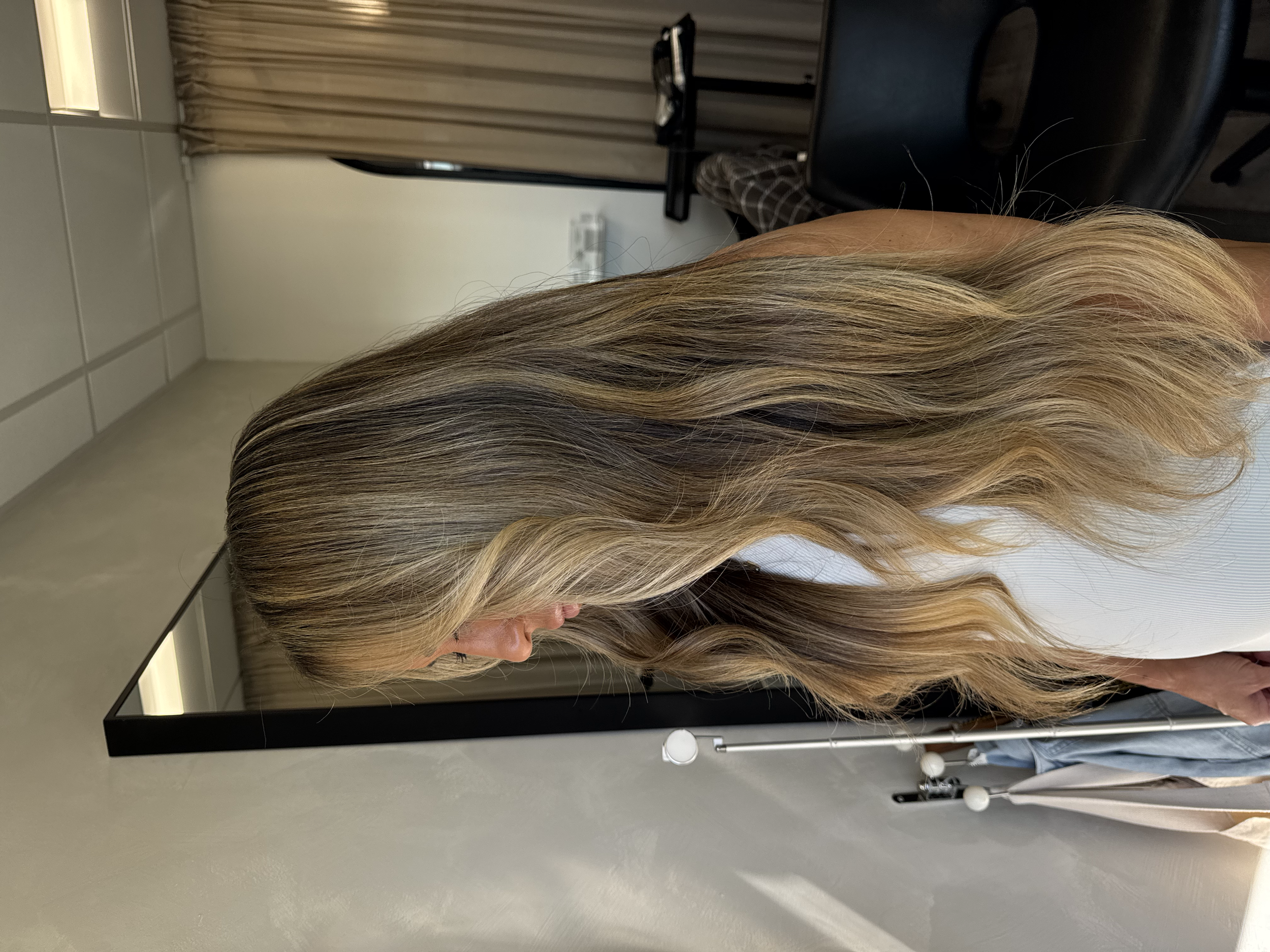 Side view of a woman with long, wavy blonde hair with darker streaks, in a room with beige curtains and gray tiled walls.