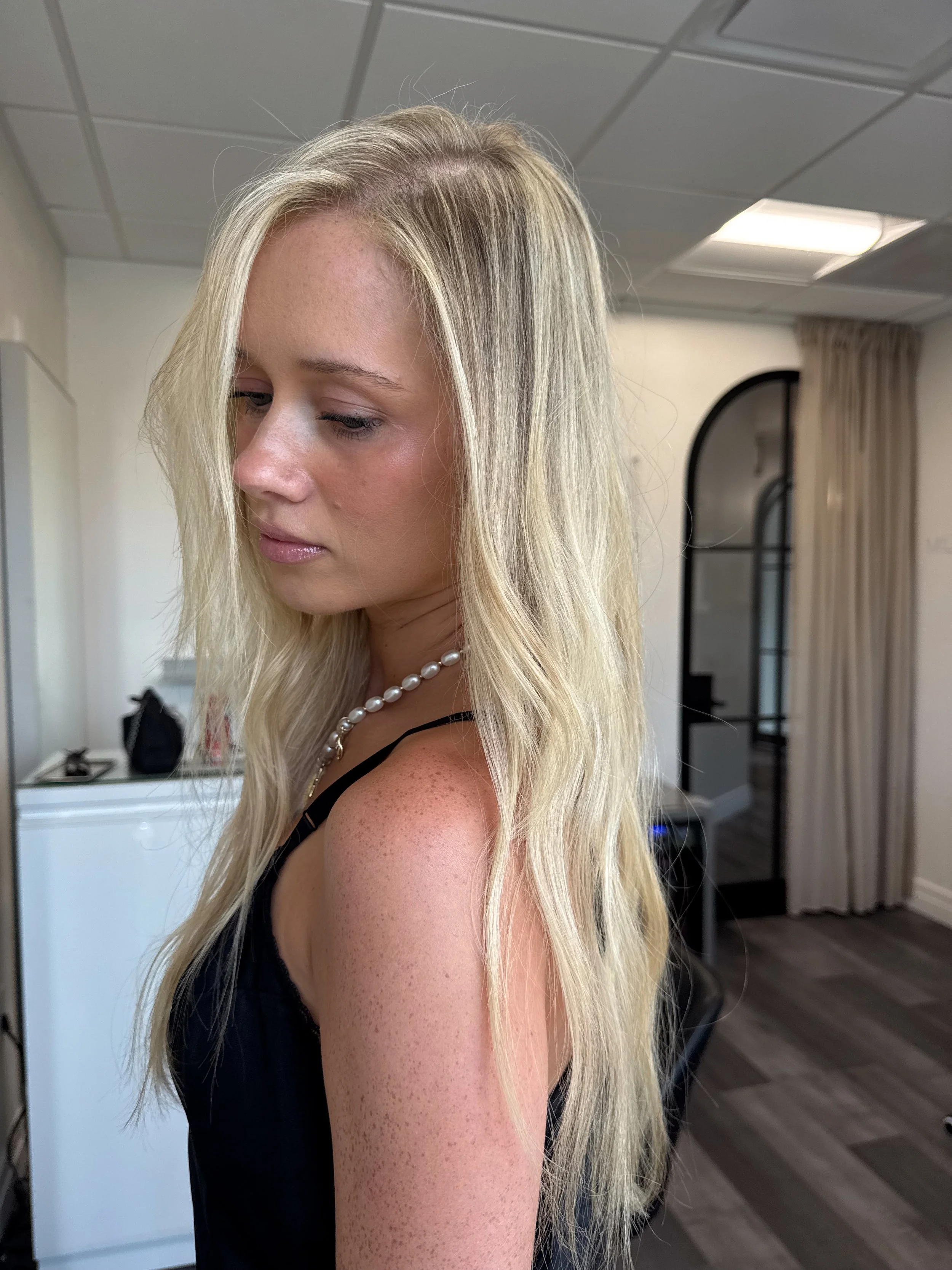 A woman with long blonde hair and freckles wearing a black sleeveless top and a pearl necklace, standing indoors near a mirror and beige curtains.