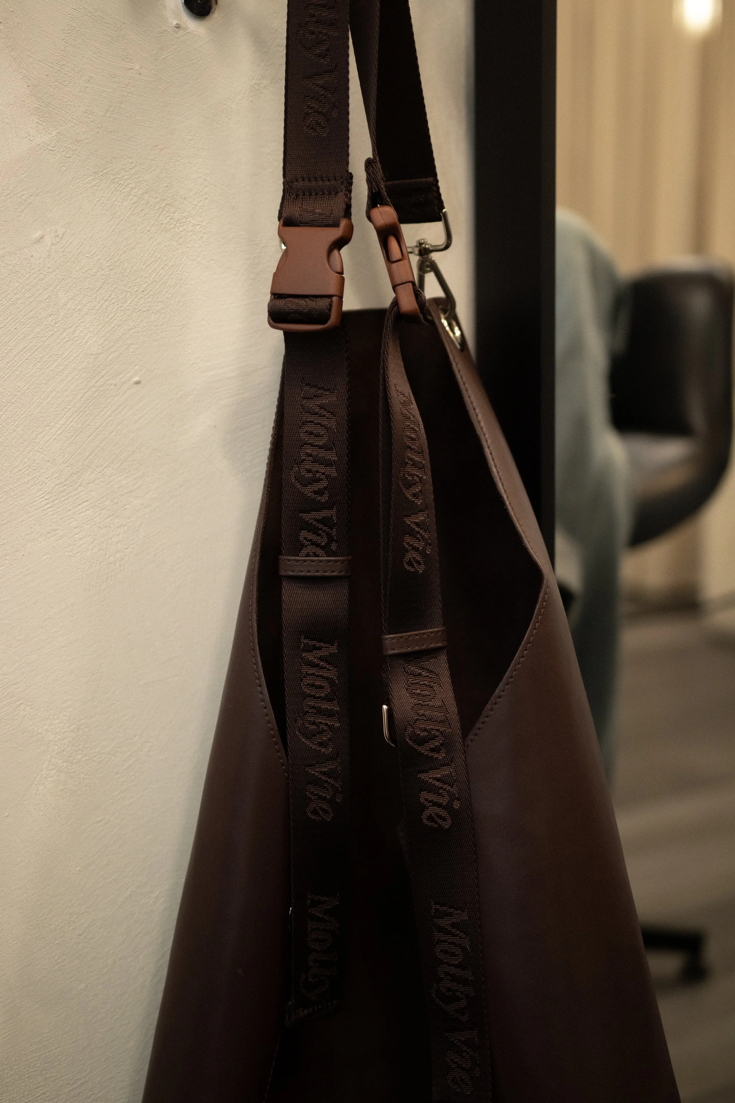 Close-up of a brown leather bag hanging on a hook, with black and brown straps, against a white textured wall. In the background, a blurred office chair is visible.