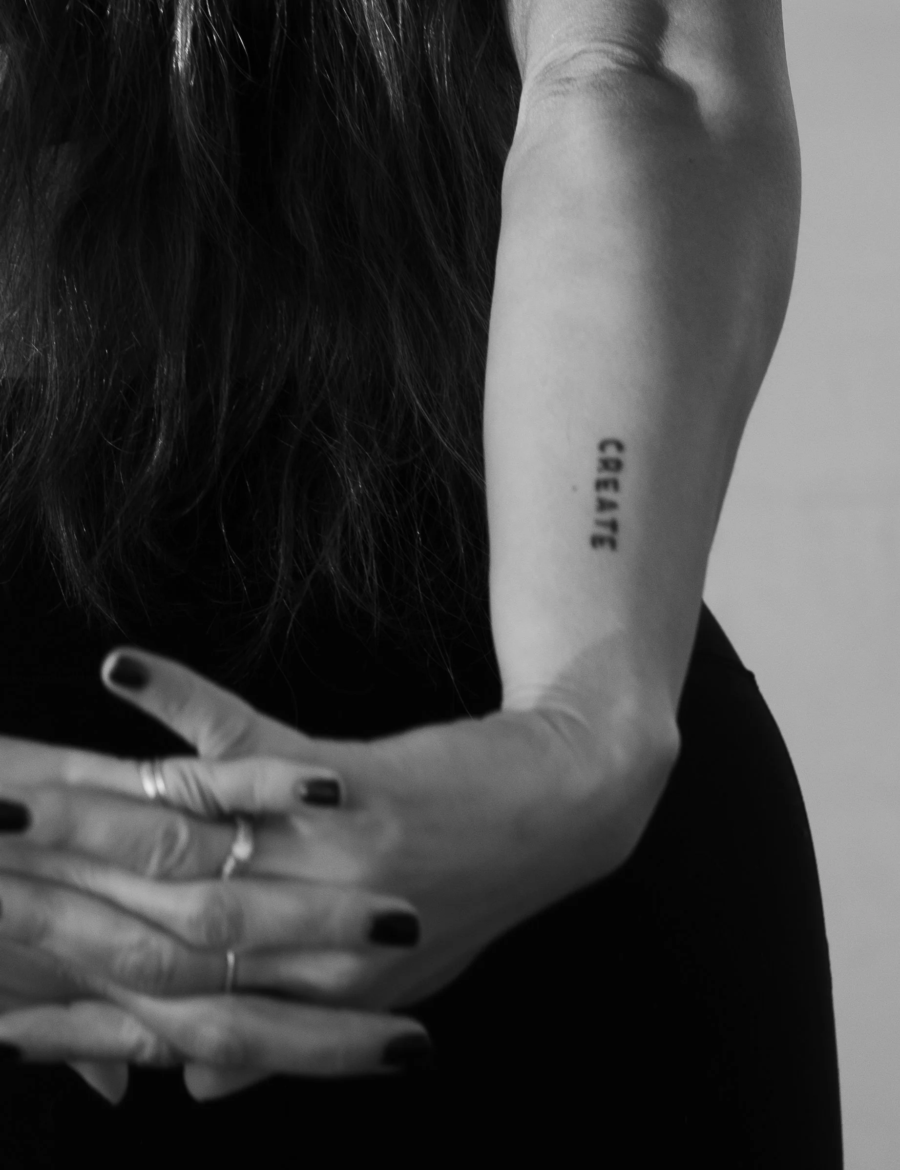 Black-and-white close-up of a person with long dark hair, black-painted fingernails, rings, and a forearm tattoo reading “CAESAR.”