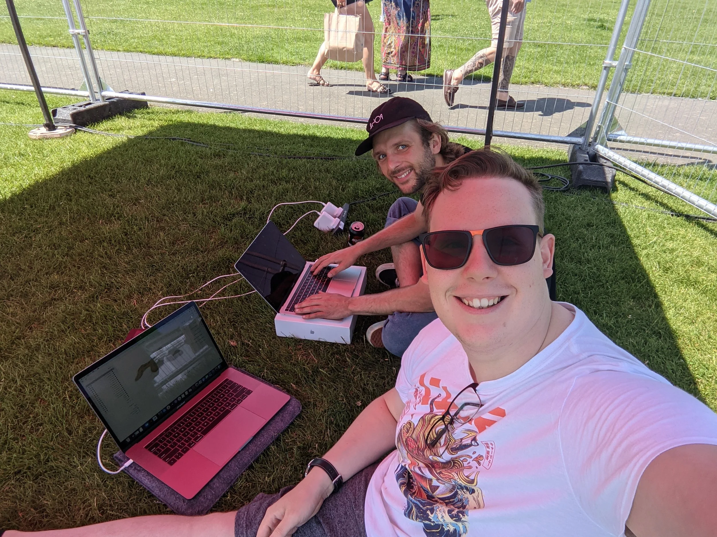 Three people are lying on the grass under a shade, with laptops and charging cables, while others walk behind a metal fence in a park on a sunny day.