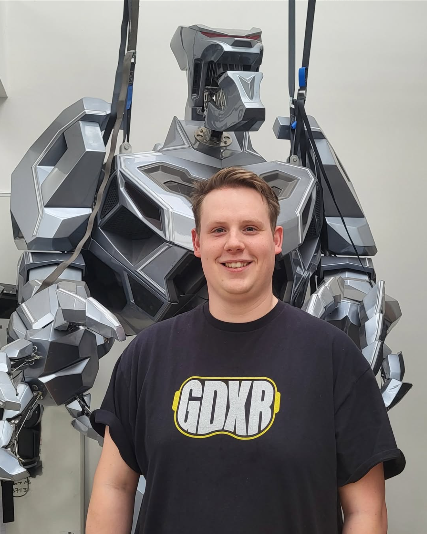 A smiling man in a black 'GDXB' t-shirt standing in front of a silver robot with a humanoid shape.