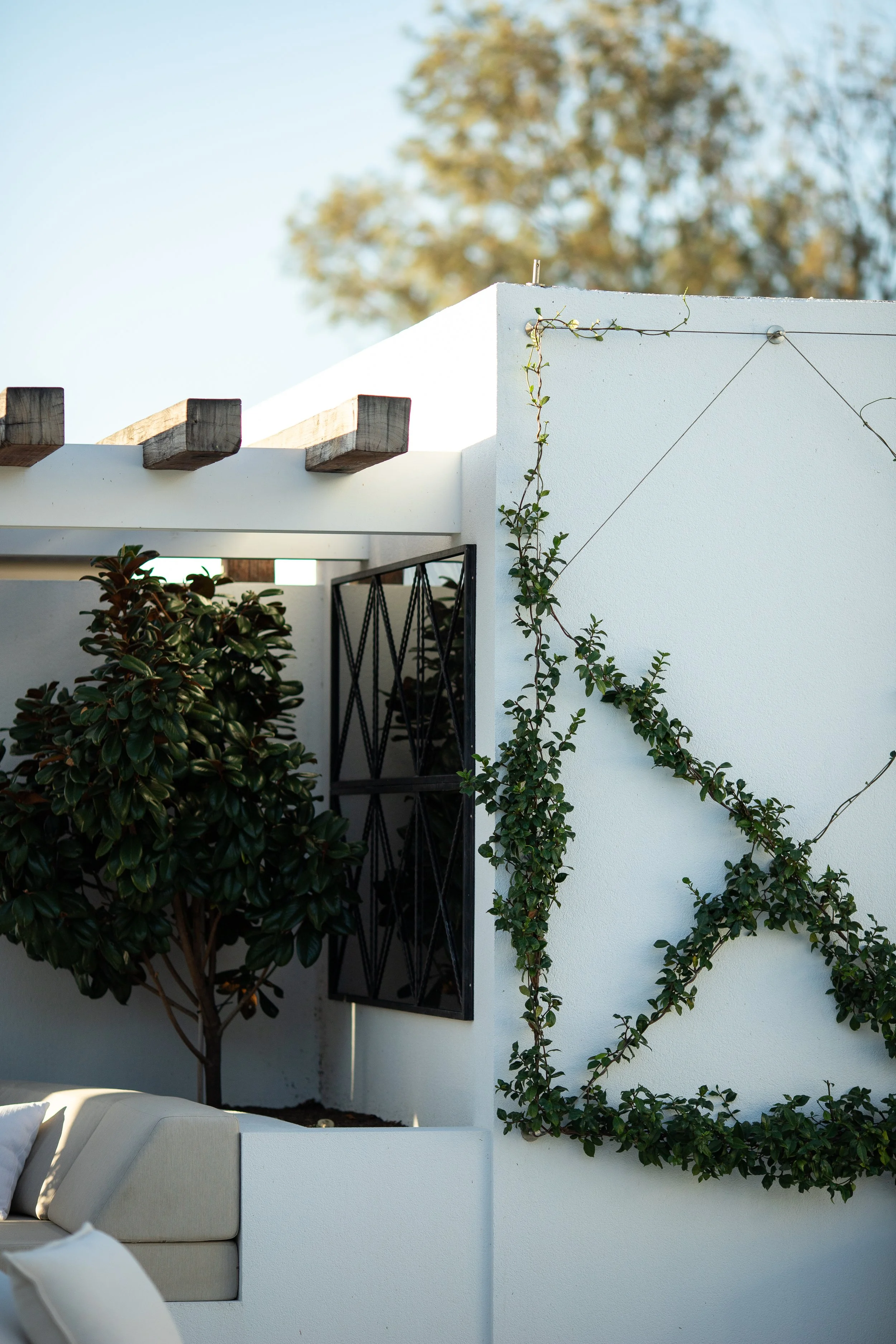 A backyard poolside patio with two modern wooden lounge chairs, a round wooden coffee table, large potted green plants, and a chandelier hanging from the ceiling. There is a white wall with louvered shutters and a fireplace in the background.