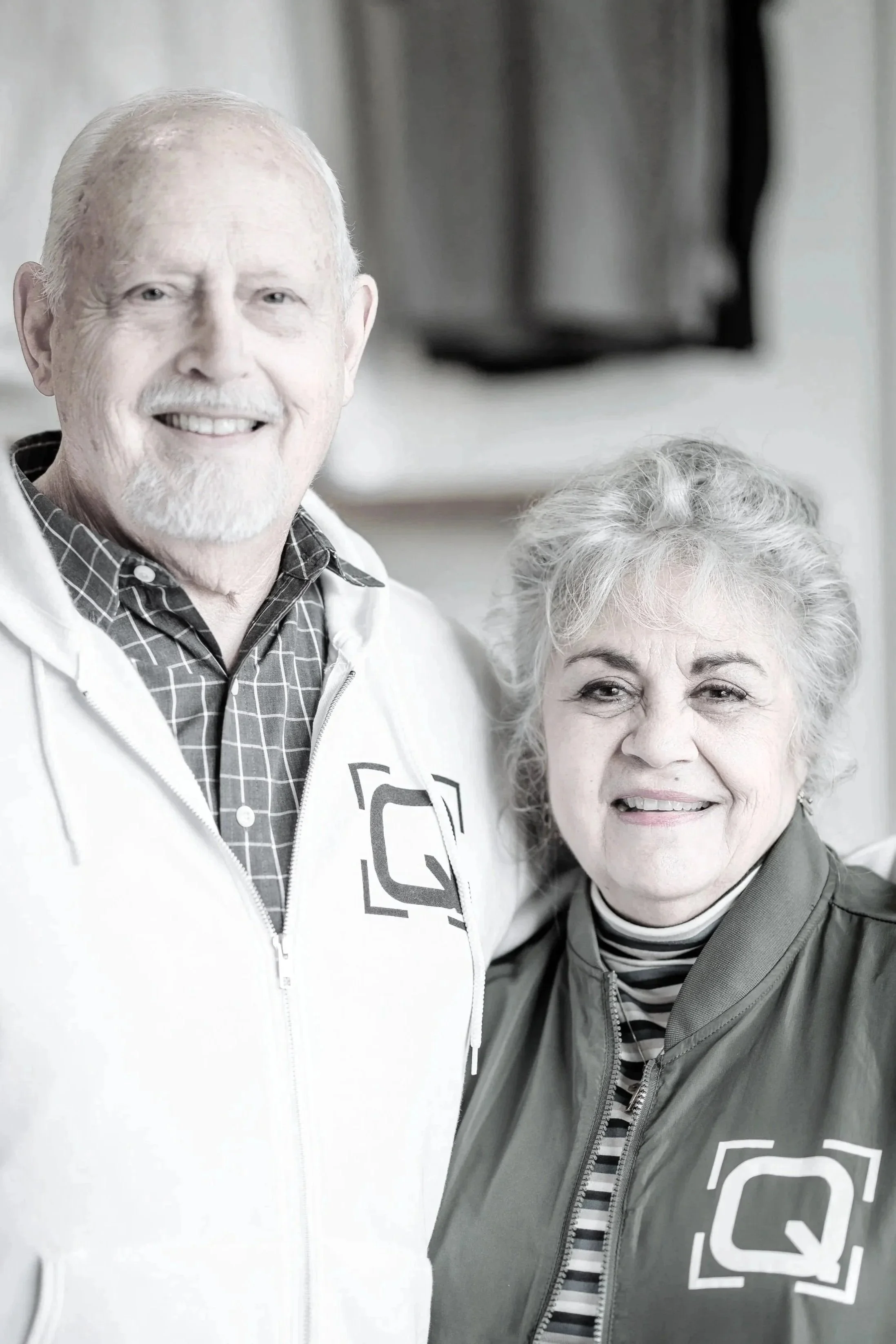 A smiling elderly man and woman standing close together indoors, both wearing jackets with a logo on them.