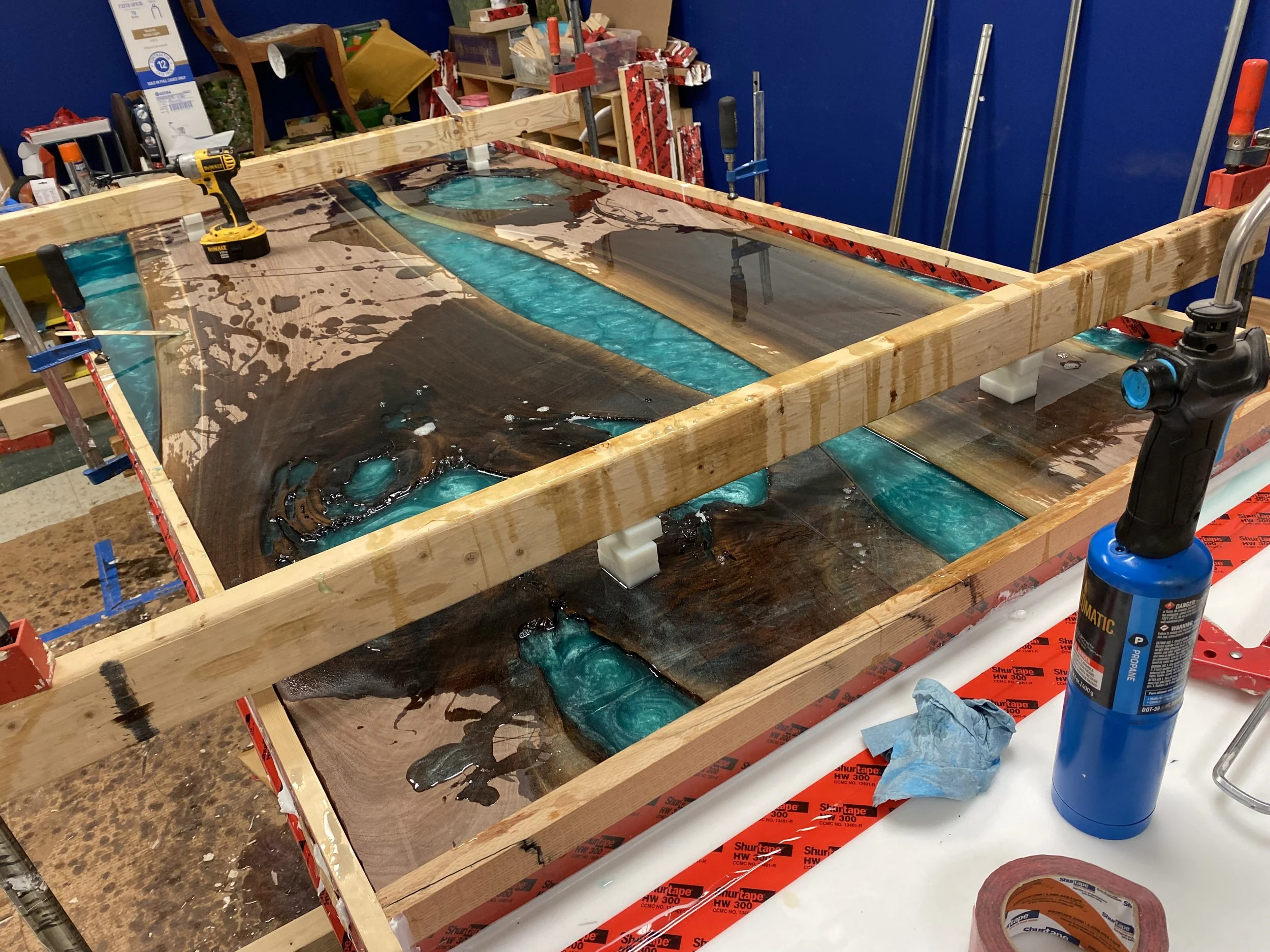 A work table with a resin river table in progress, featuring a wooden frame, resin poured to create a river effect, and tools like a drill and a blow torch nearby.