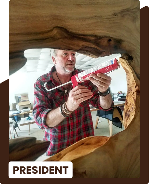 A man wearing a red plaid shirt applying caulk or sealant from a tube with a caulk gun, seen through a hollowed-out wooden frame, in an office or workshop setting.