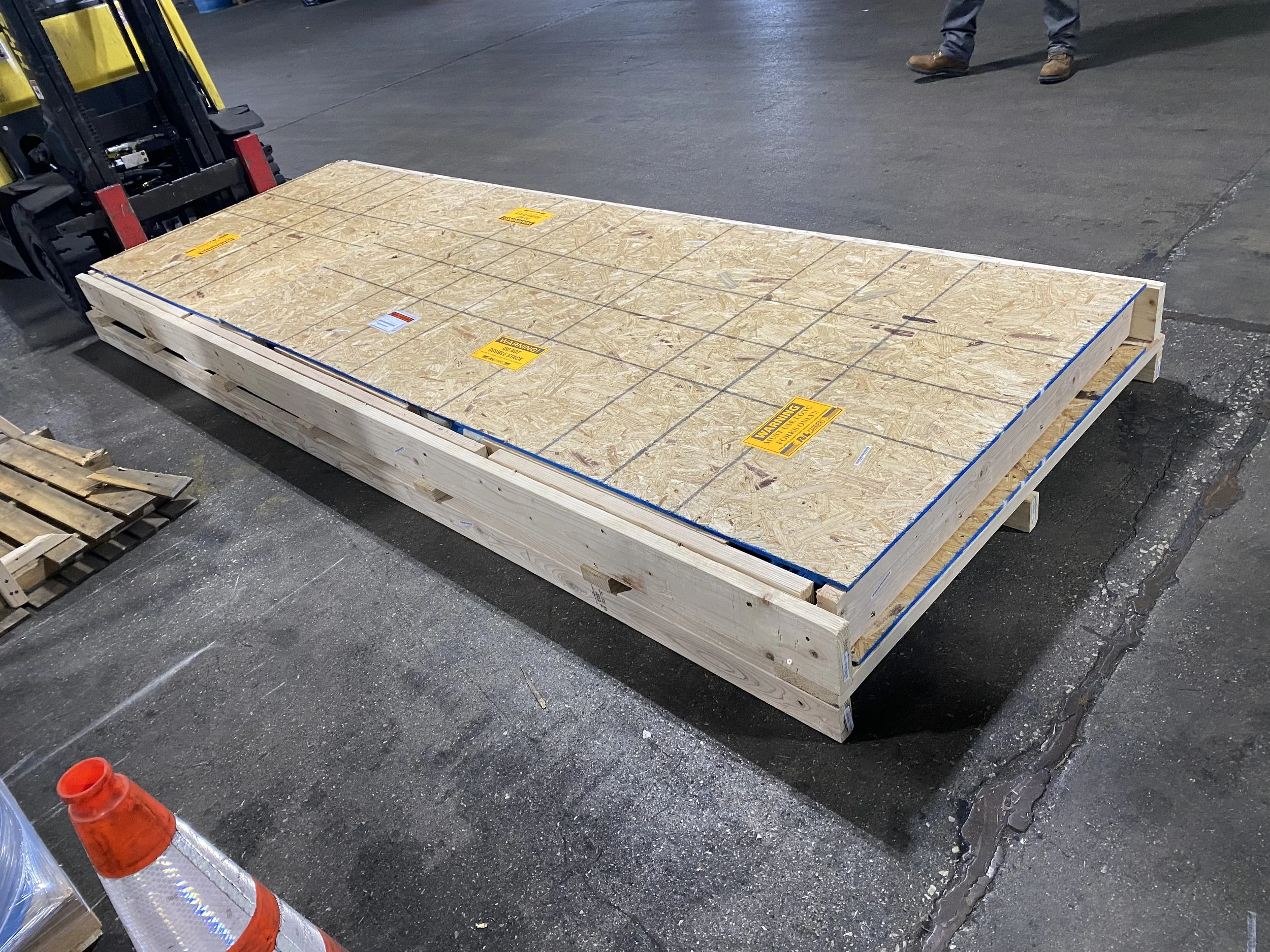 Large wooden crate with warning labels and a forklift nearby in an industrial warehouse setting.