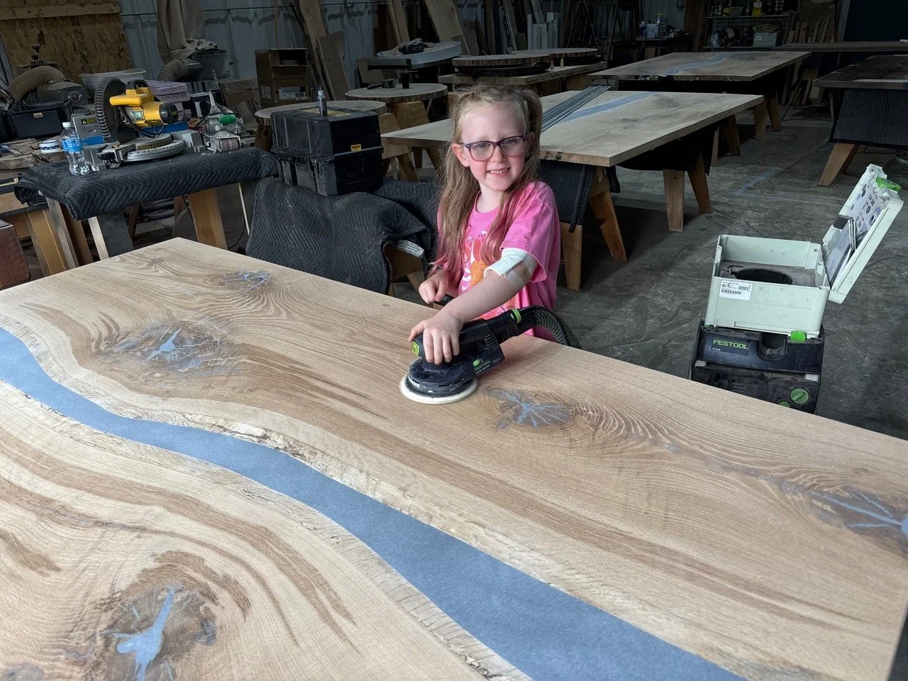 A young girl with glasses and a pink shirt is sanding a large wooden slab with a handheld rotary sander in a woodworking shop. She is smiling while working.
