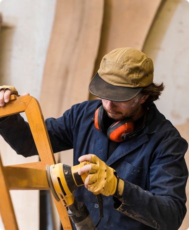 Man wearing a cap and protective gear, using a drill on a wooden chair in a workshop.