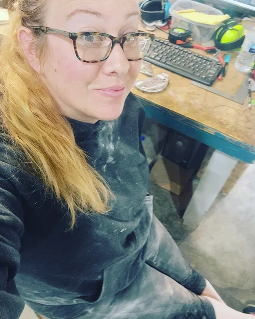 A woman with glasses and long red hair sitting in a workshop with a cluttered workbench in the background.