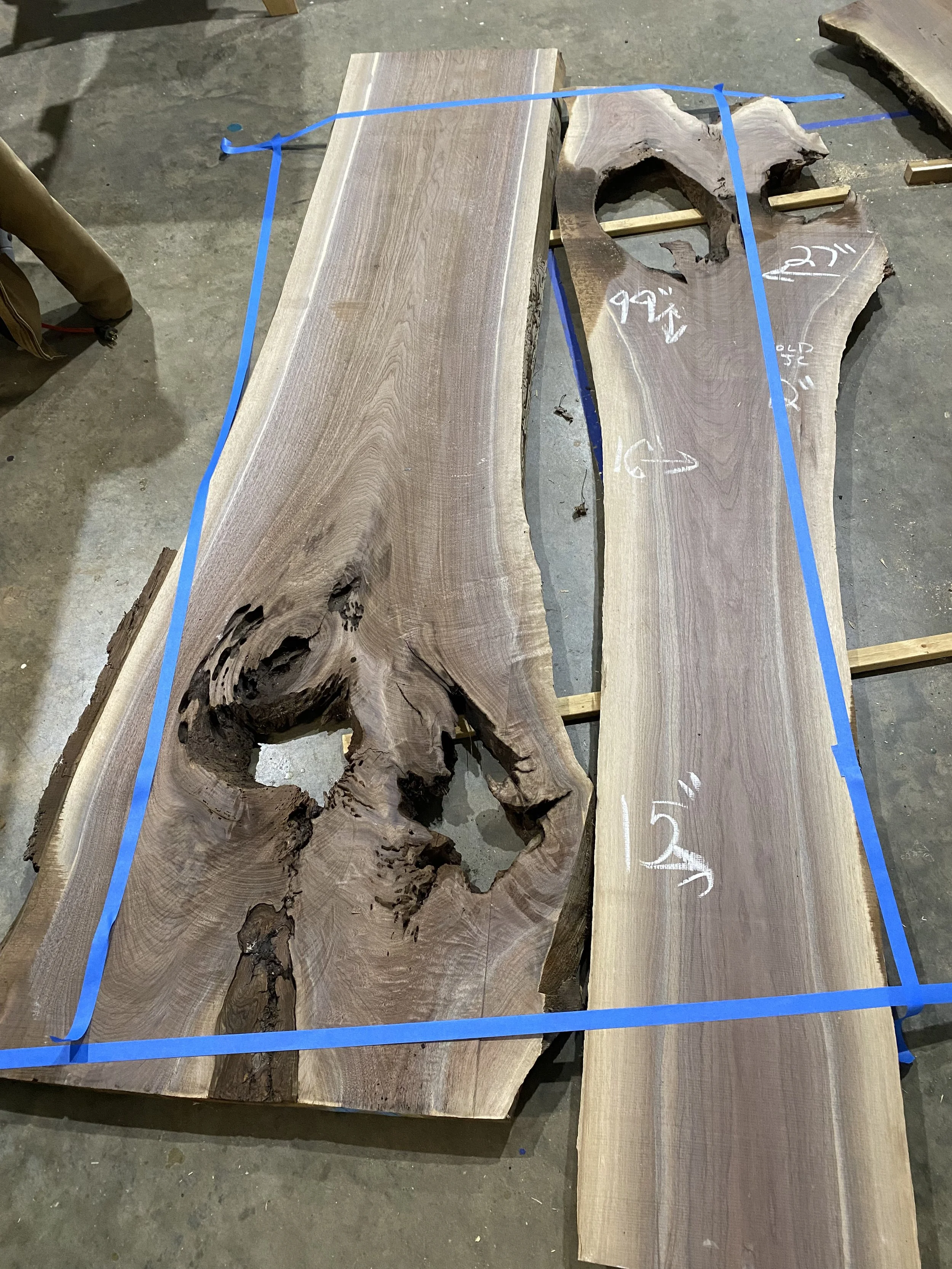 Unfinished live edge wooden slabs with natural knots and cracks, measured and marked, laid on a concrete floor, blue painter's tape outlining the edges.