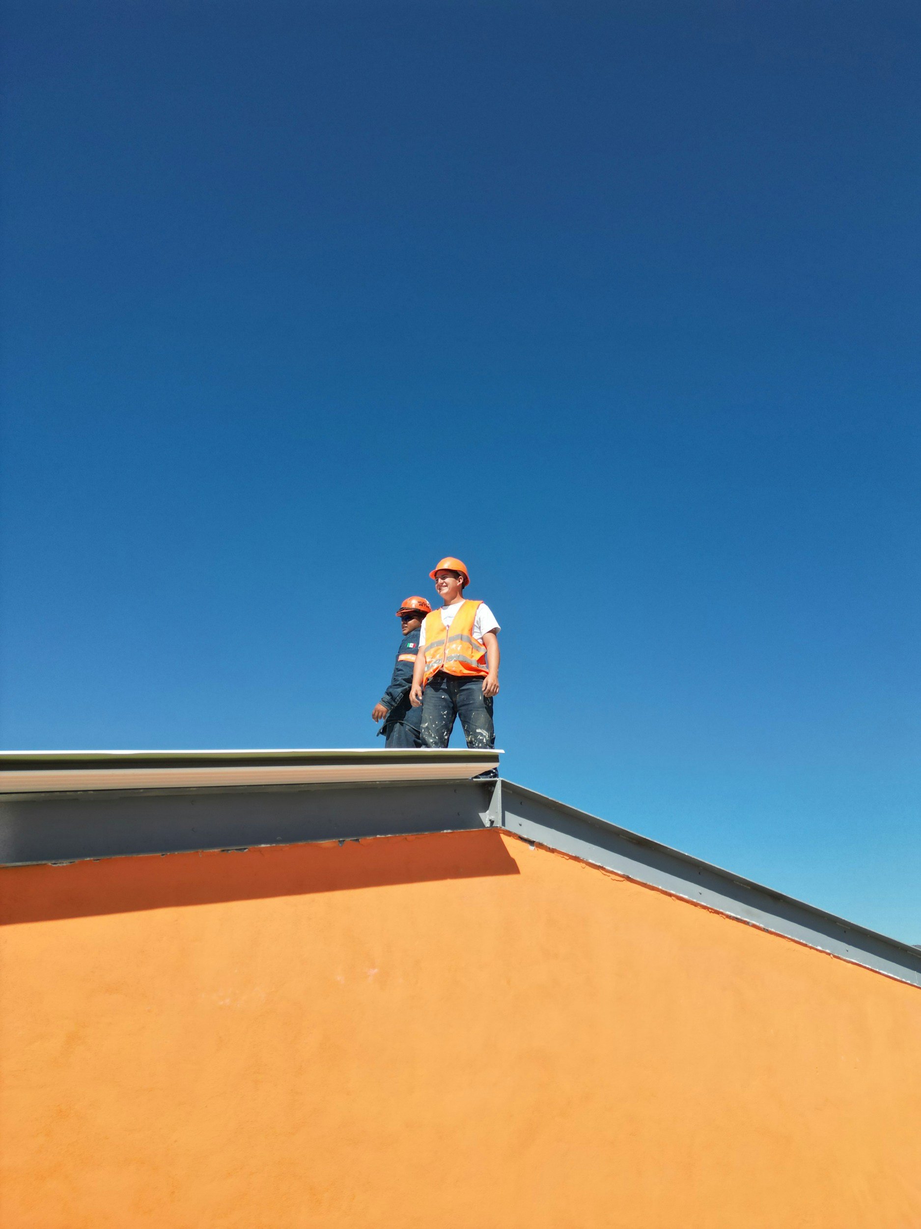 Two construction workers wearing orange hats and safety vests standing on the roof of a building against a blue sky.