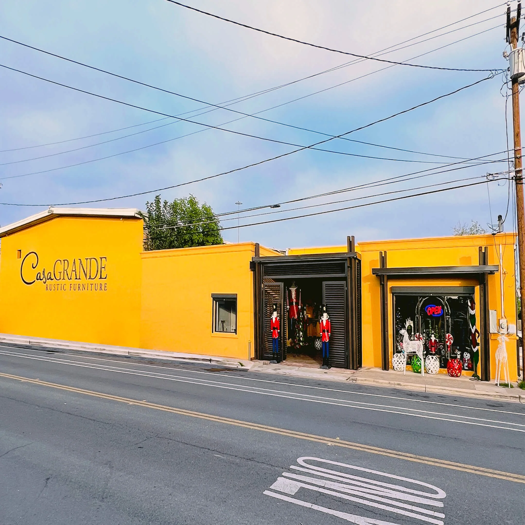 Yellow building with black accents displaying 'Casa GRANDE RUSTIC FURNITURE' sign, with store entrance and window decorations, on a street with crosswalk and utility wires overhead.