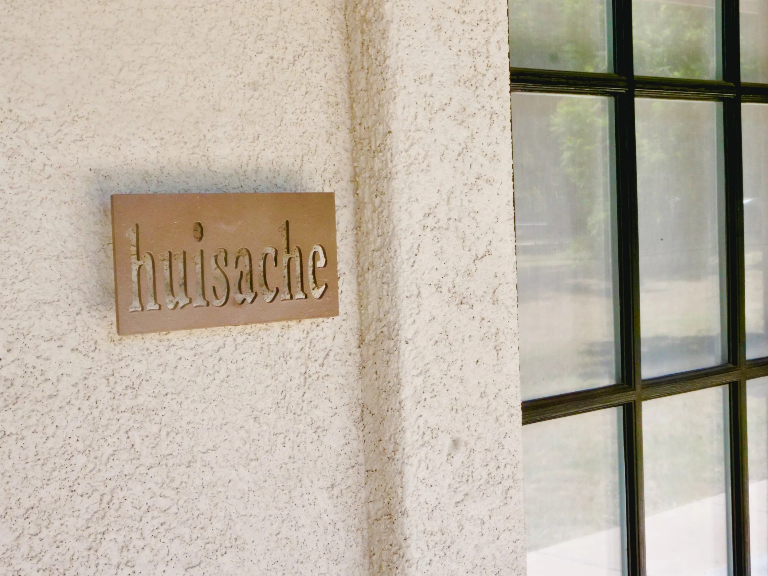 Close-up of a beige wall with a brown sign that reads 'hüsache' and a nearby window with multiple panes and black framing.