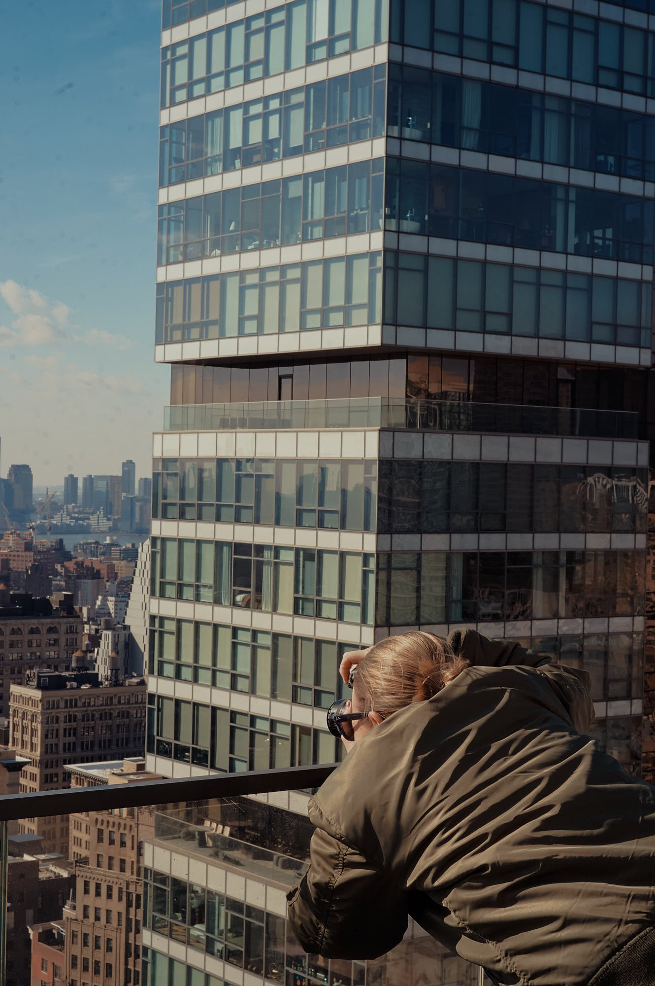 A person in a green jacket with brown hair and sunglasses leaning on a balcony railing, taking a photo of a tall glass building with a city skyline in the background.