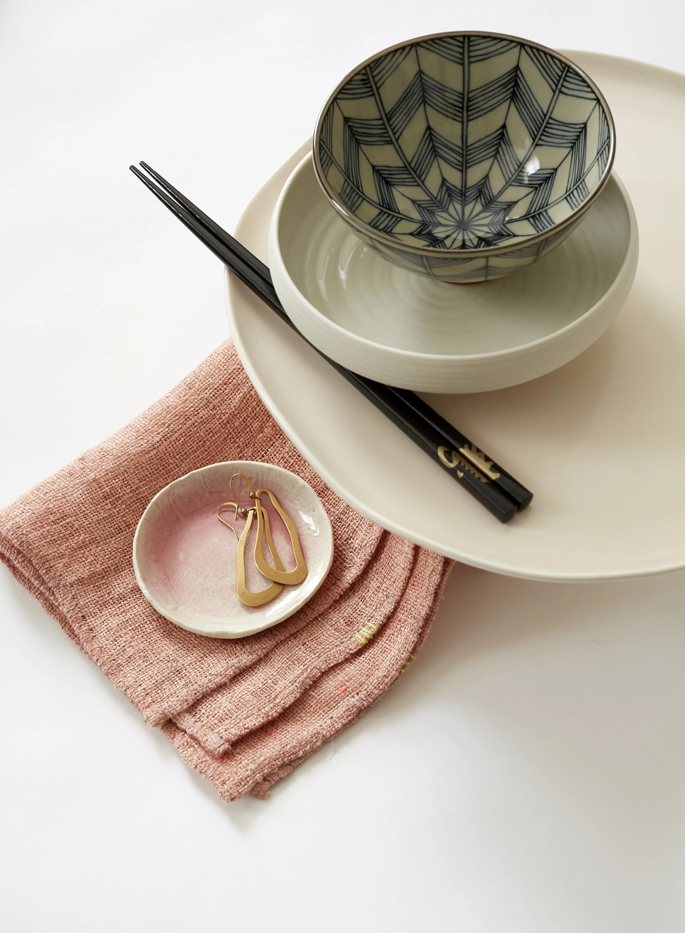 Flat lay of stacked plates, a bowl with a geometric leaf pattern, a small pink dish with gold earrings, pink cloth, and black chopsticks.