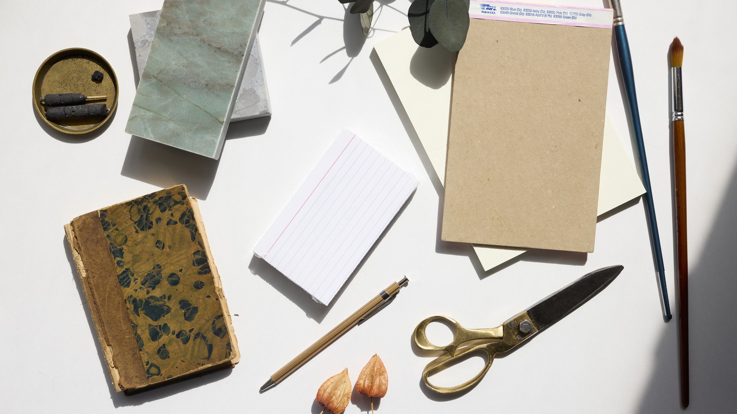 Flat lay of various stationery items on a white surface, including notebooks, paper, scissors, pens, and small decorative objects.