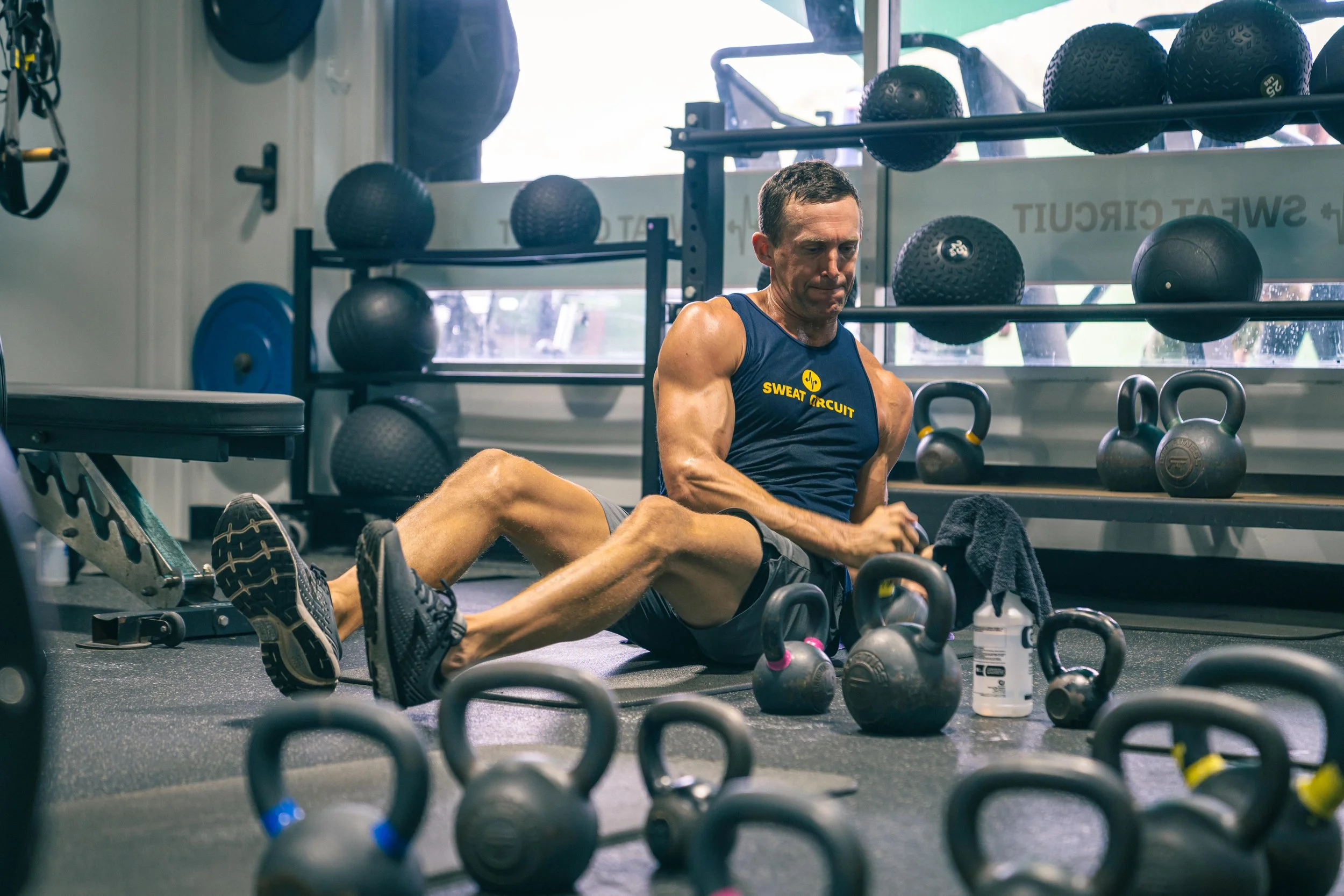 A man in a blue tank top with 'SWEAT CIRCUIT' printed on it, performing a sit-up on the floor of a gym surrounded by black kettlebells and exercise balls, with gym equipment and a large window in the background.