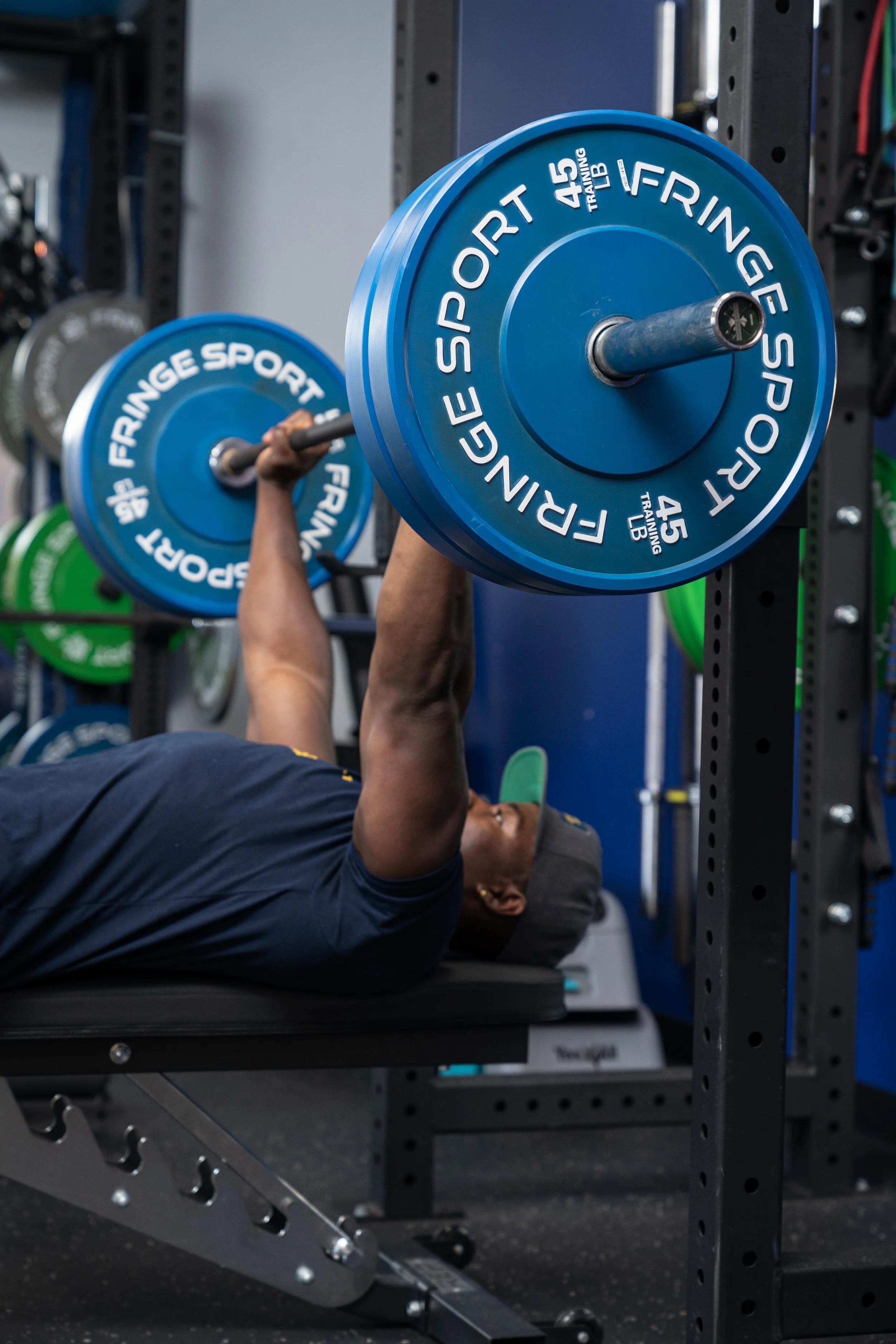 Person lying on a workout bench lifting a large blue weight plate with the words 'FREIGE SPORT 45' and 'TRAILING 45 LB' on it.