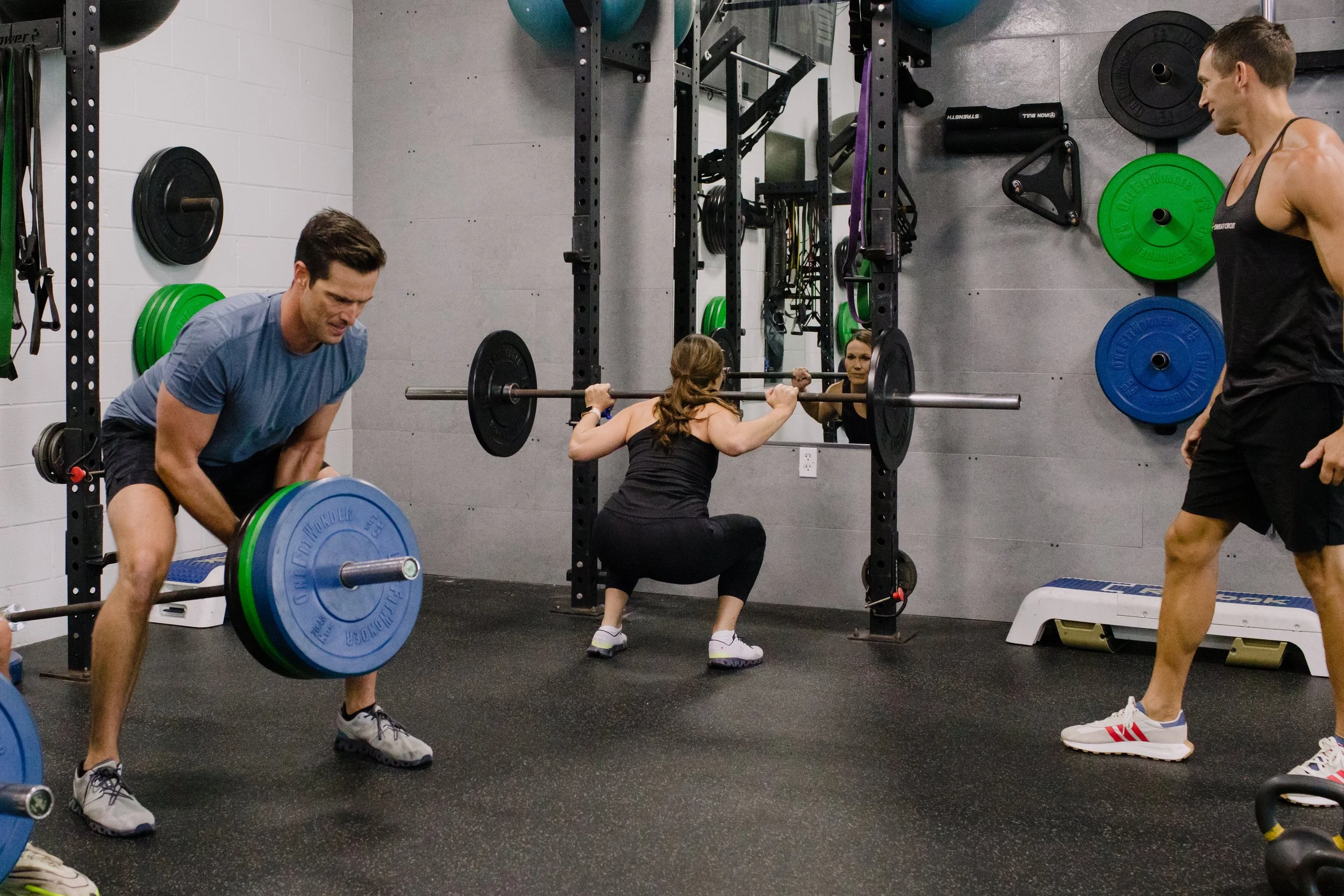 People working out in a gym. A woman is performing a squat with a barbell, a man is lifting a barbell, and another person is standing nearby. Gym equipment, weights, and mirrors are visible.