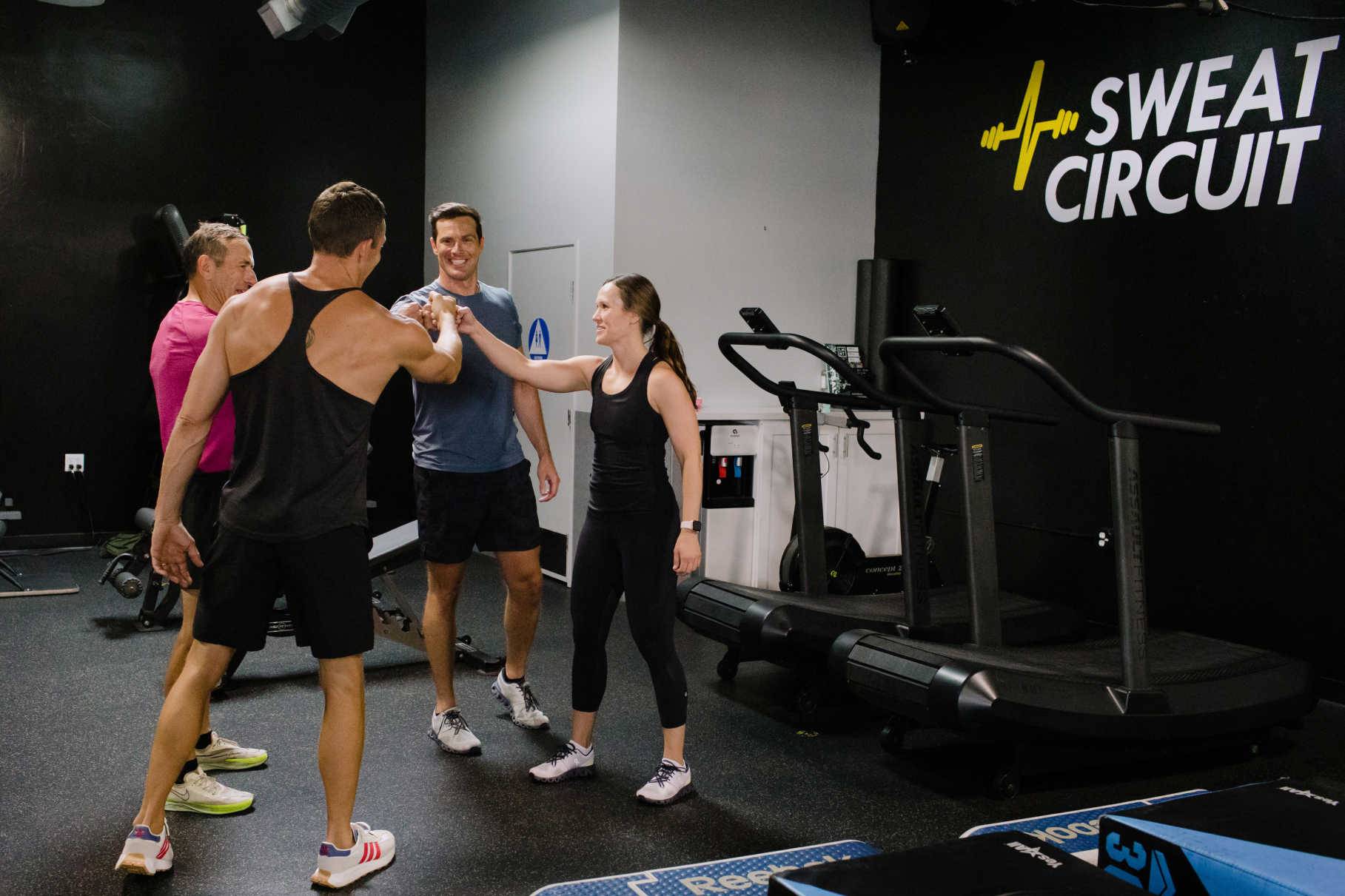 Group of five people inside a gym, four men and one woman, celebrating and smiling. One man is giving a fist bump to the woman. The gym has fitness equipment including treadmills and weights. The logo 'SWEAT CIRCUIT' is visible on the wall.