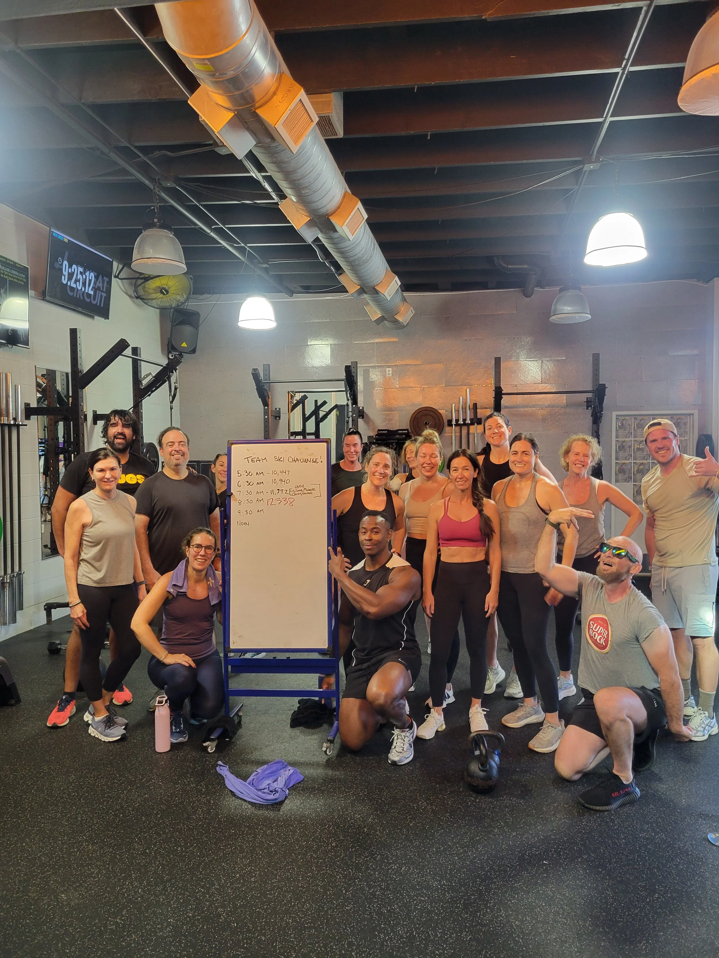 A group of people in athletic clothing after a workout at the gym, smiling and posing together in front of a whiteboard with a schedule for a team ski challenge, inside a gym with black flooring, lighting fixtures, and gym equipment in the background