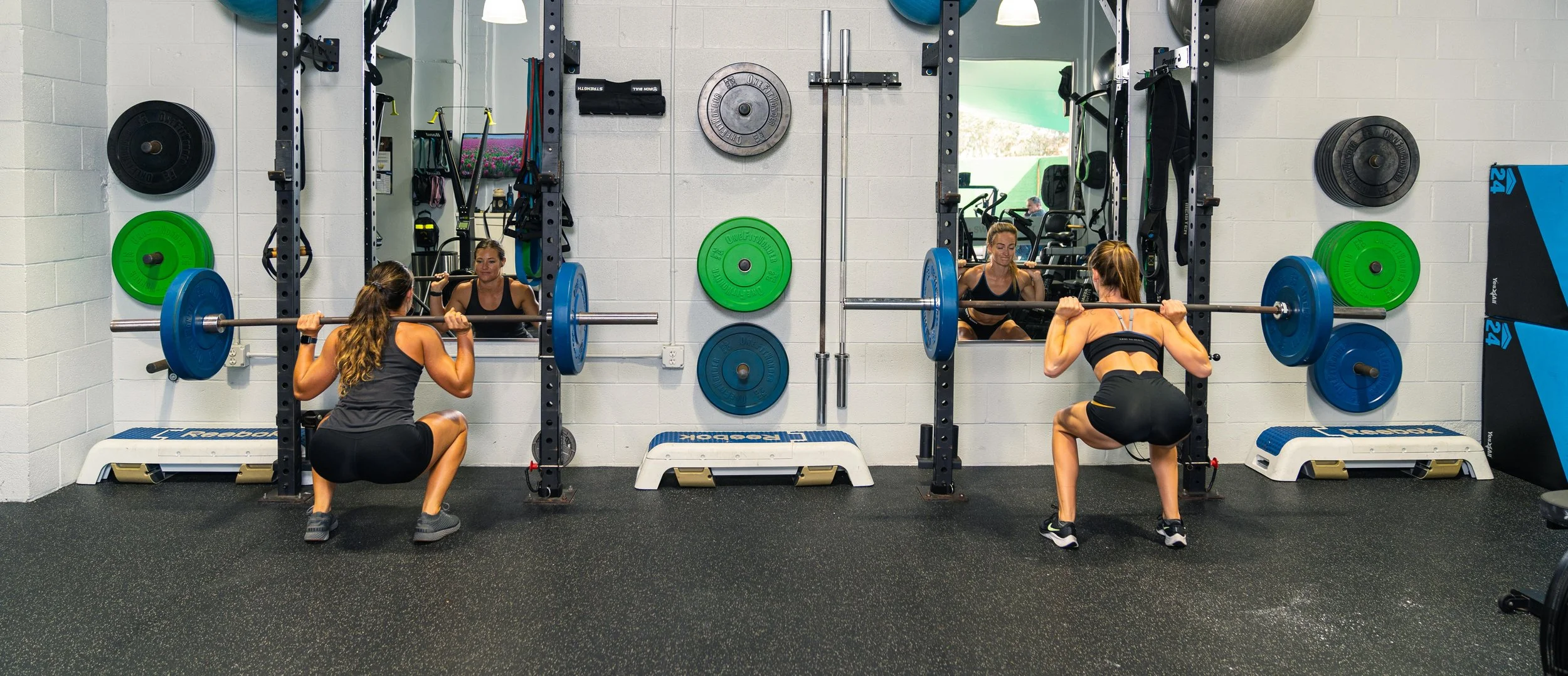 Two women squat with barbells loaded with blue weight plates in a gym, seen from the back. They are in front of a mirror, with various colored weight plates on the wall and gym equipment visible in the background.