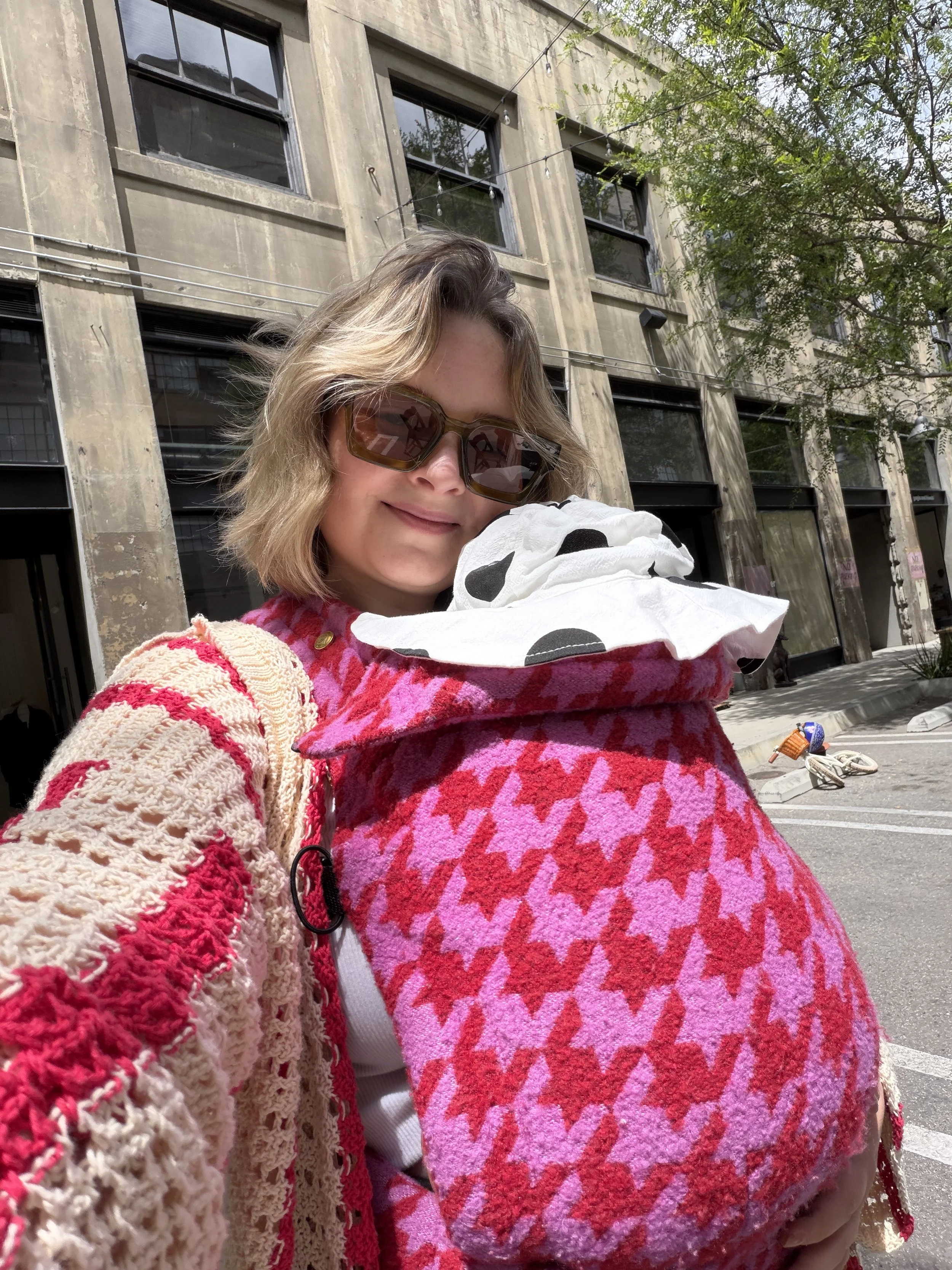 A woman with short blonde hair wearing sunglasses, a colorful pink and red patterned baby carrier, and holding a baby with a black polka dot hat. She is smiling and standing outdoors in front of a building with large windows and trees.