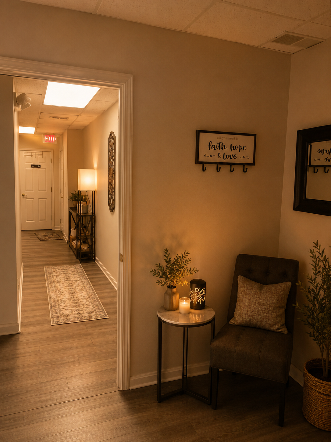 Cozy indoor space with a small side table holding candles and a plant, a comfortable chair with pillow, wall decor with the words 'faith, hope, & love,' and a mirror reflecting a well-lit hallway with a rug and door.