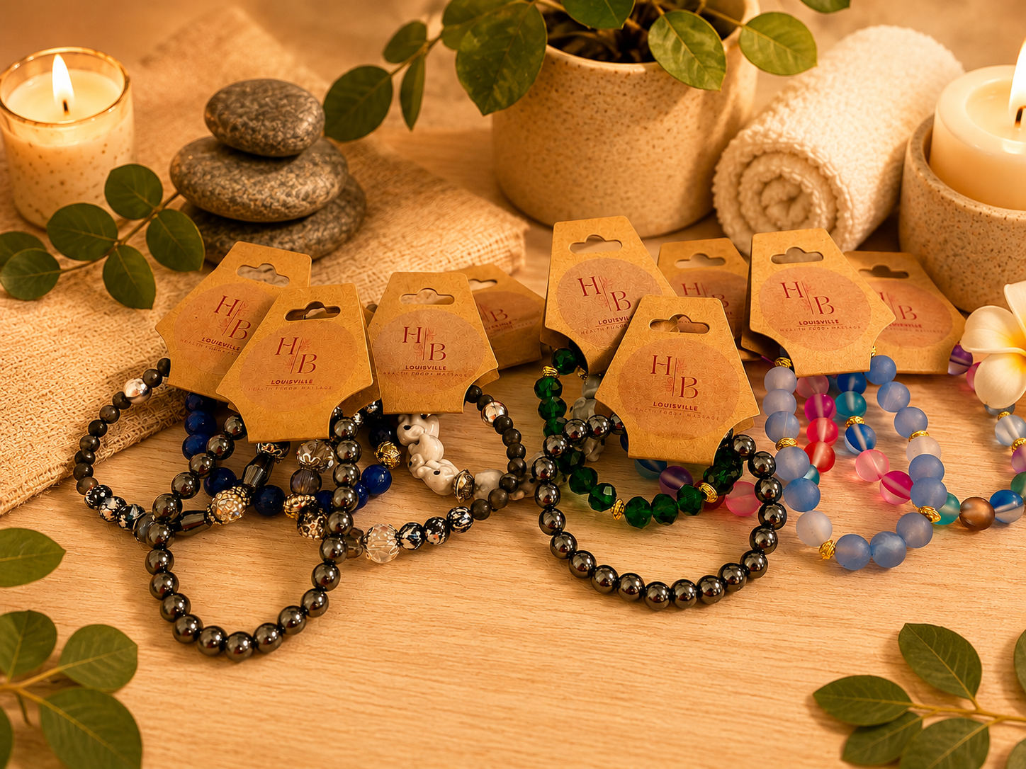 Colorful beaded bracelets with tags reading "H B Louisville" displayed on a wooden surface surrounded by candles, rolled towels, green leaves, and a potted plant, creating a spa-like or decorative setting.