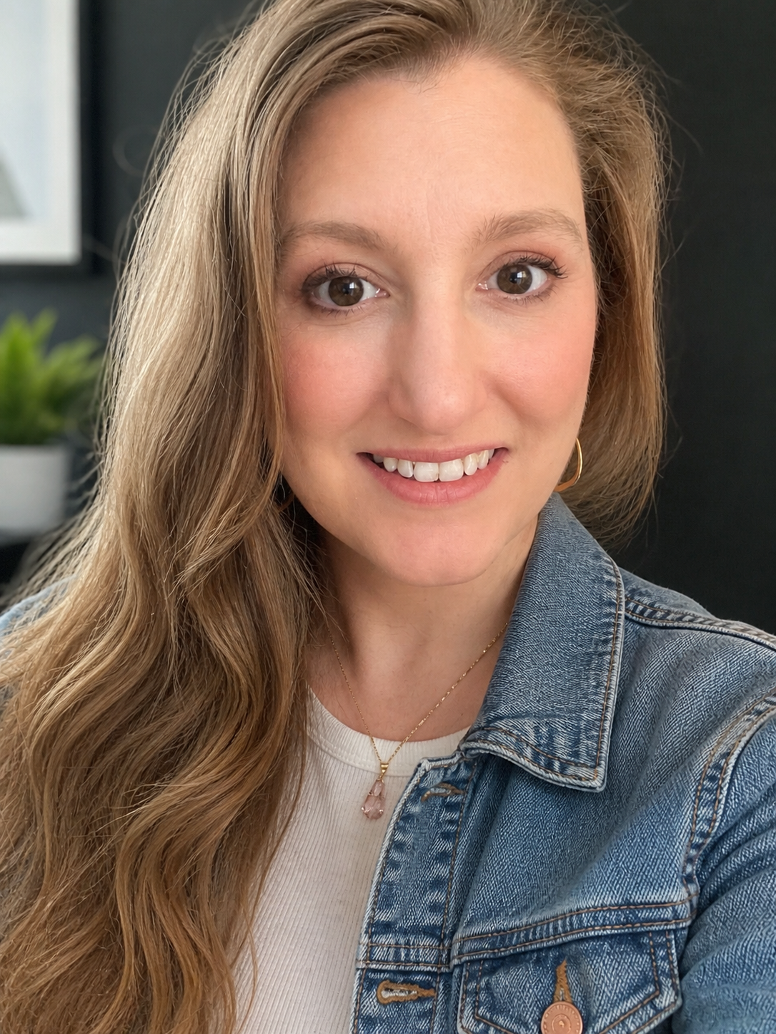 A woman with wavy, long blonde hair, wearing a denim jacket, a white top, and gold jewelry, smiling at the camera.
