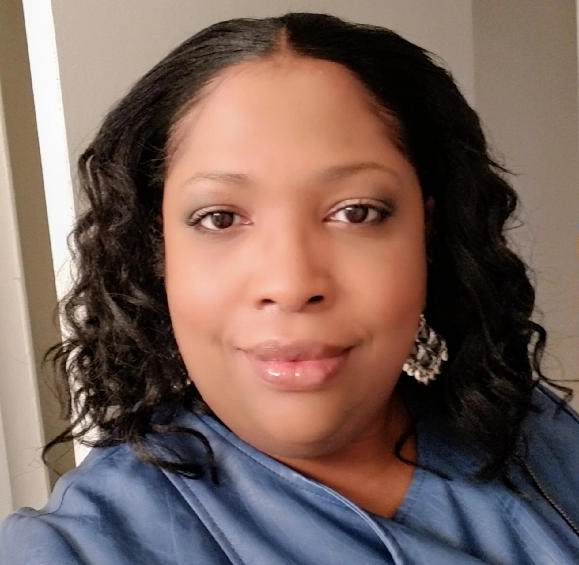 Close-up photo of a woman with curly black hair, wearing a blue shirt, in a room with beige walls.
