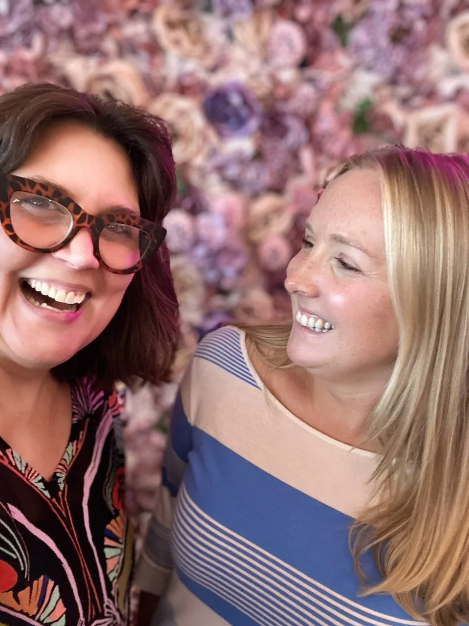 Two women smiling at each other in front of a floral background.
