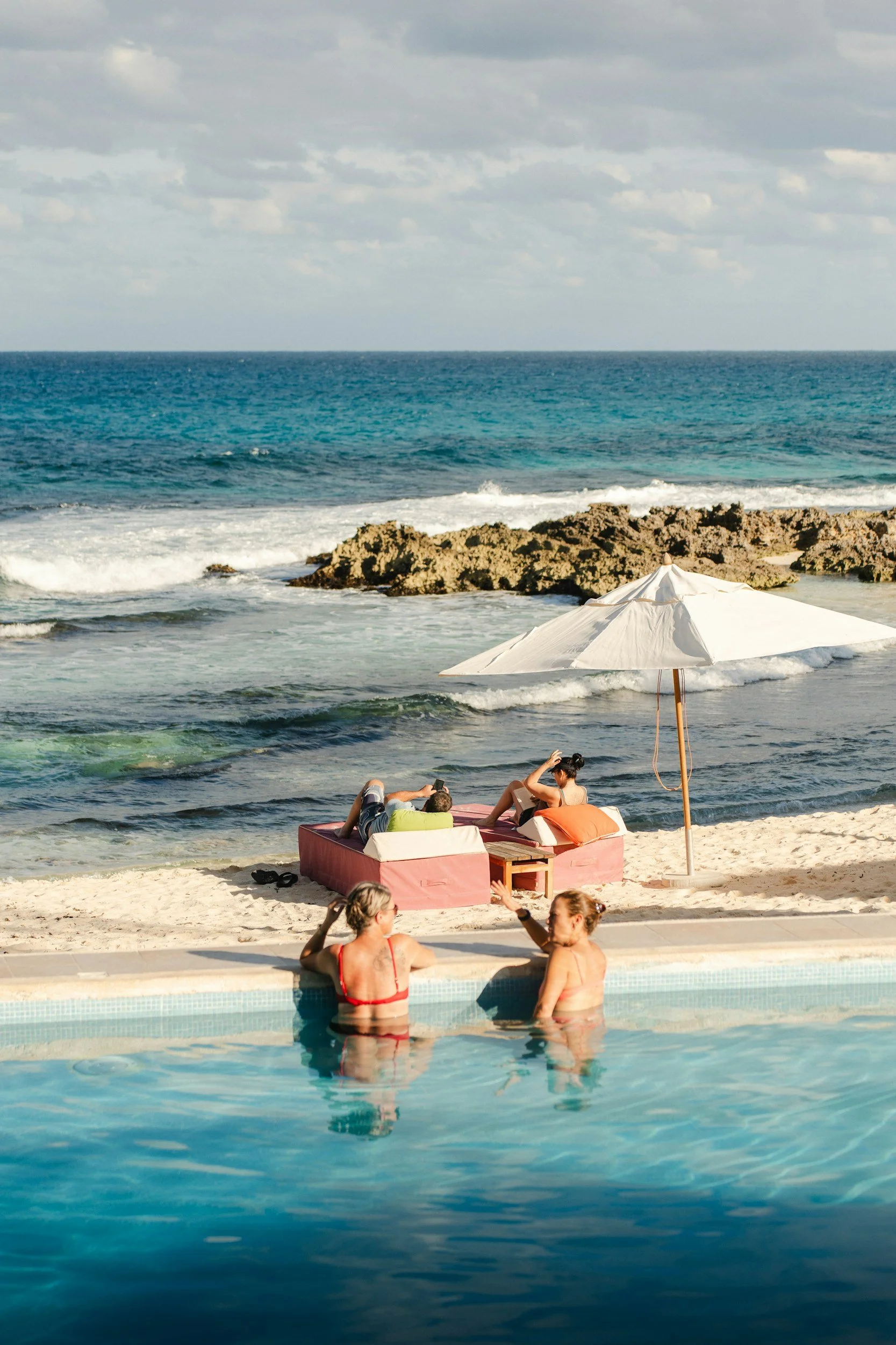 People relaxing at a beachside pool with ocean view, lounge chairs, and an umbrella.