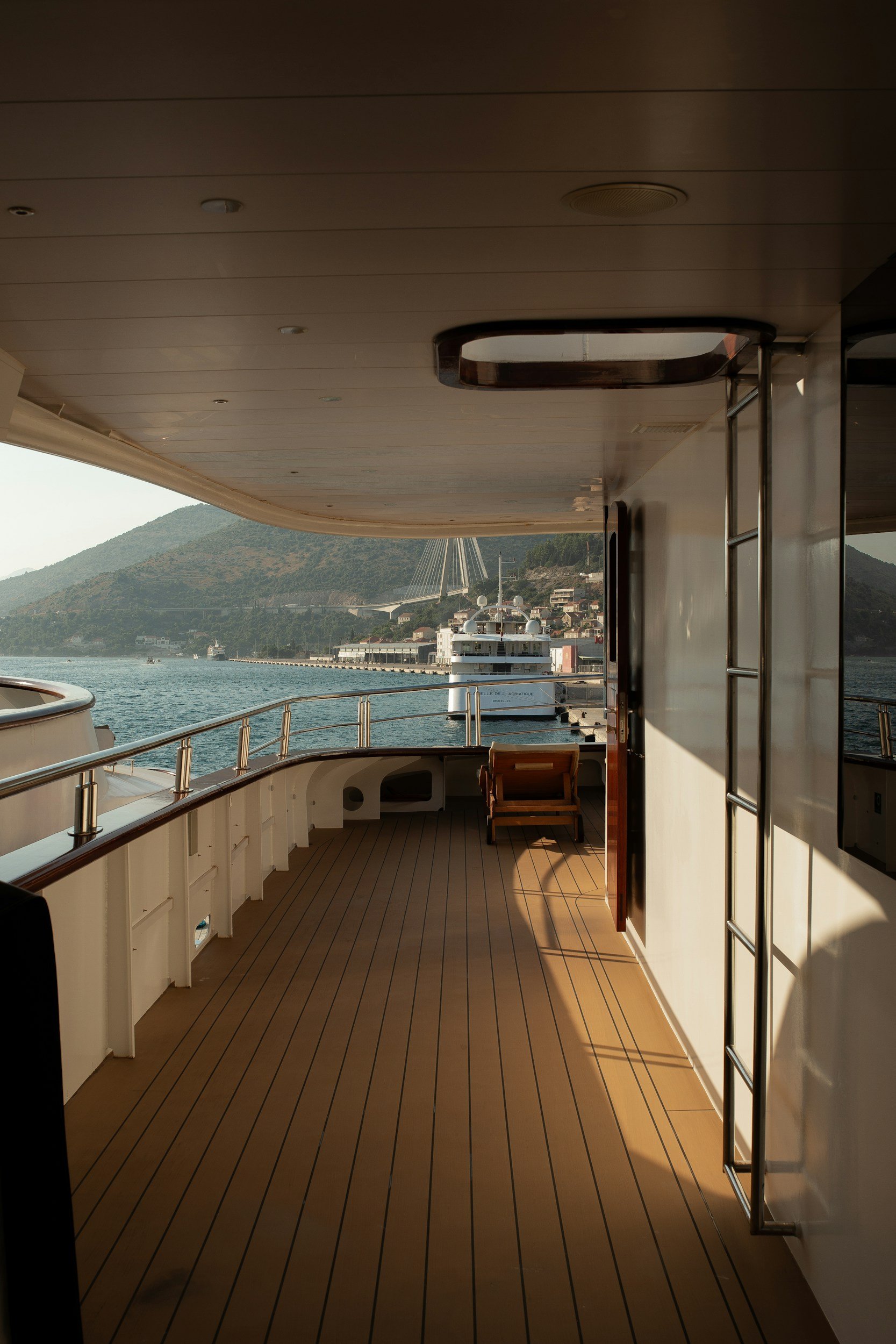 View from a yacht deck showing water, mountains, and another yacht in harbor with a bridge and buildings in the background.
