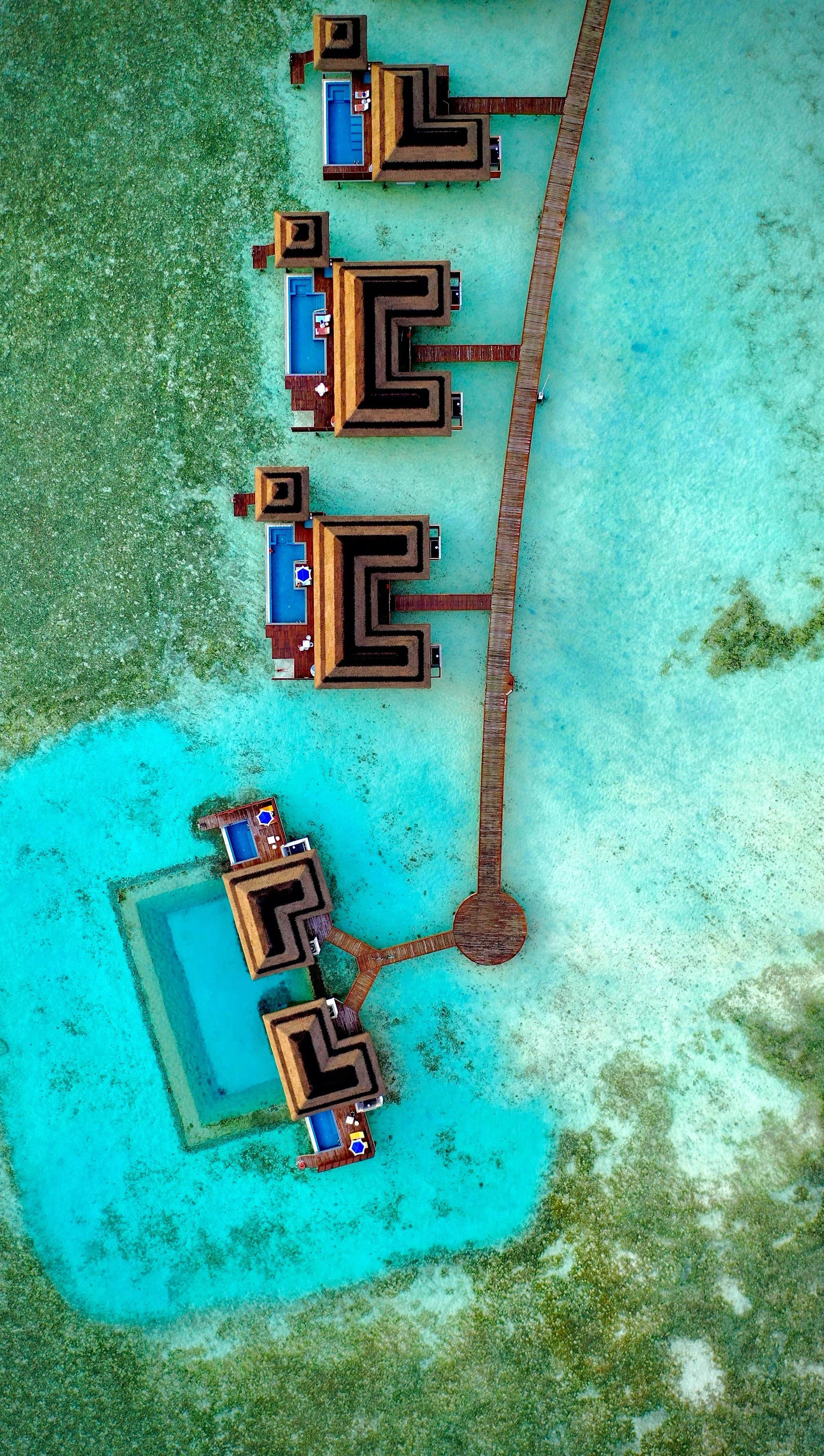 Aerial view of a resort with multiple wooden bungalows with blue pools, connected by a wooden walkway, surrounded by water and greenery.