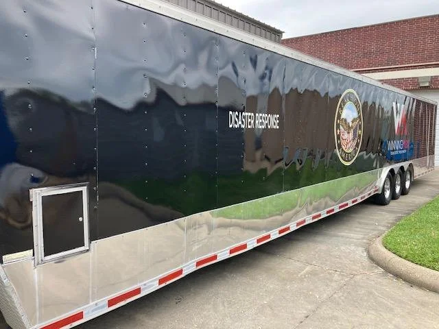 A large black disaster response trailer with official logos and text, parked on a concrete surface near a brick building and grassy area.