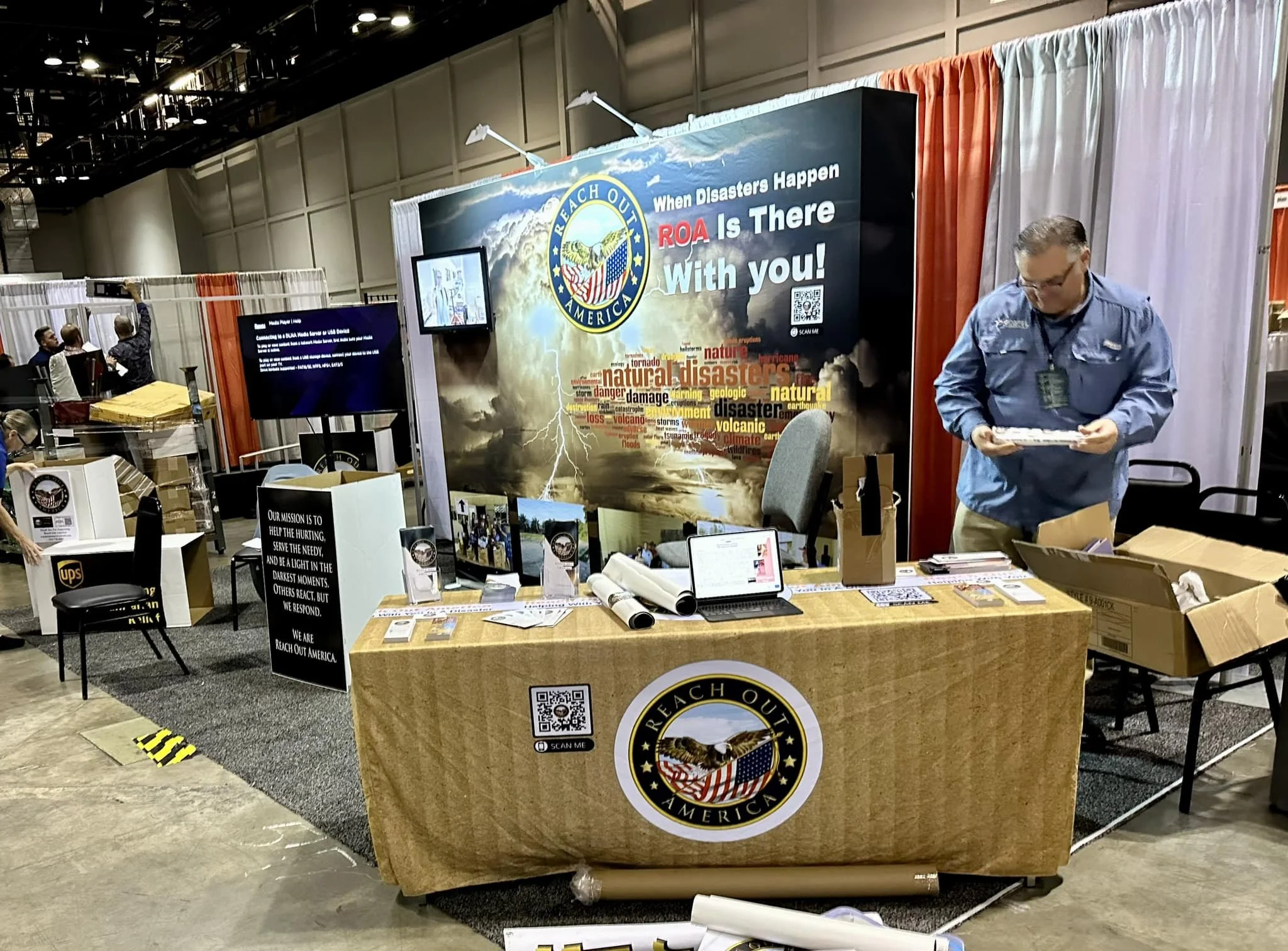 An exhibition booth for Reach Out America featuring a large backdrop with storm imagery and text about natural disasters. The booth has a wooden table with a Reach Out America logo, informational flyers, a tablet, and a person organizing materials. T