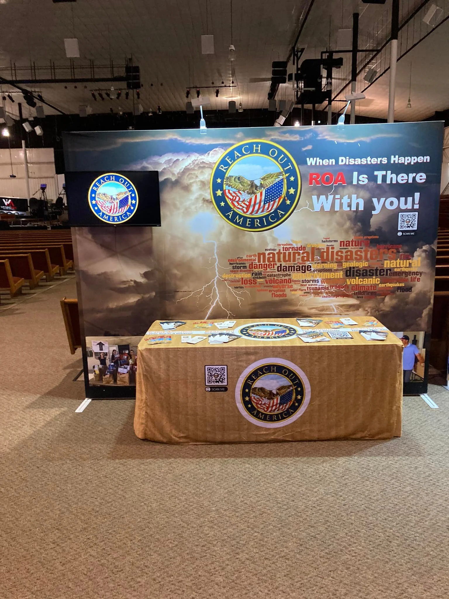 An informational booth with a large banner about natural disasters and emergency preparedness by Reach Out America, featuring a thunderstorm graphic, a lightning bolt, and words related to disasters. There is a table with flyers and QR codes, set in 