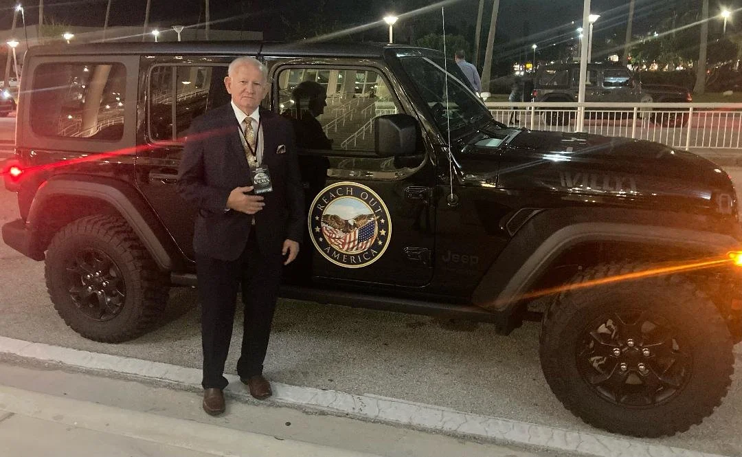 A man in a suit standing next to a black Jeep with a 'Reach Out America' emblem on the door at night.