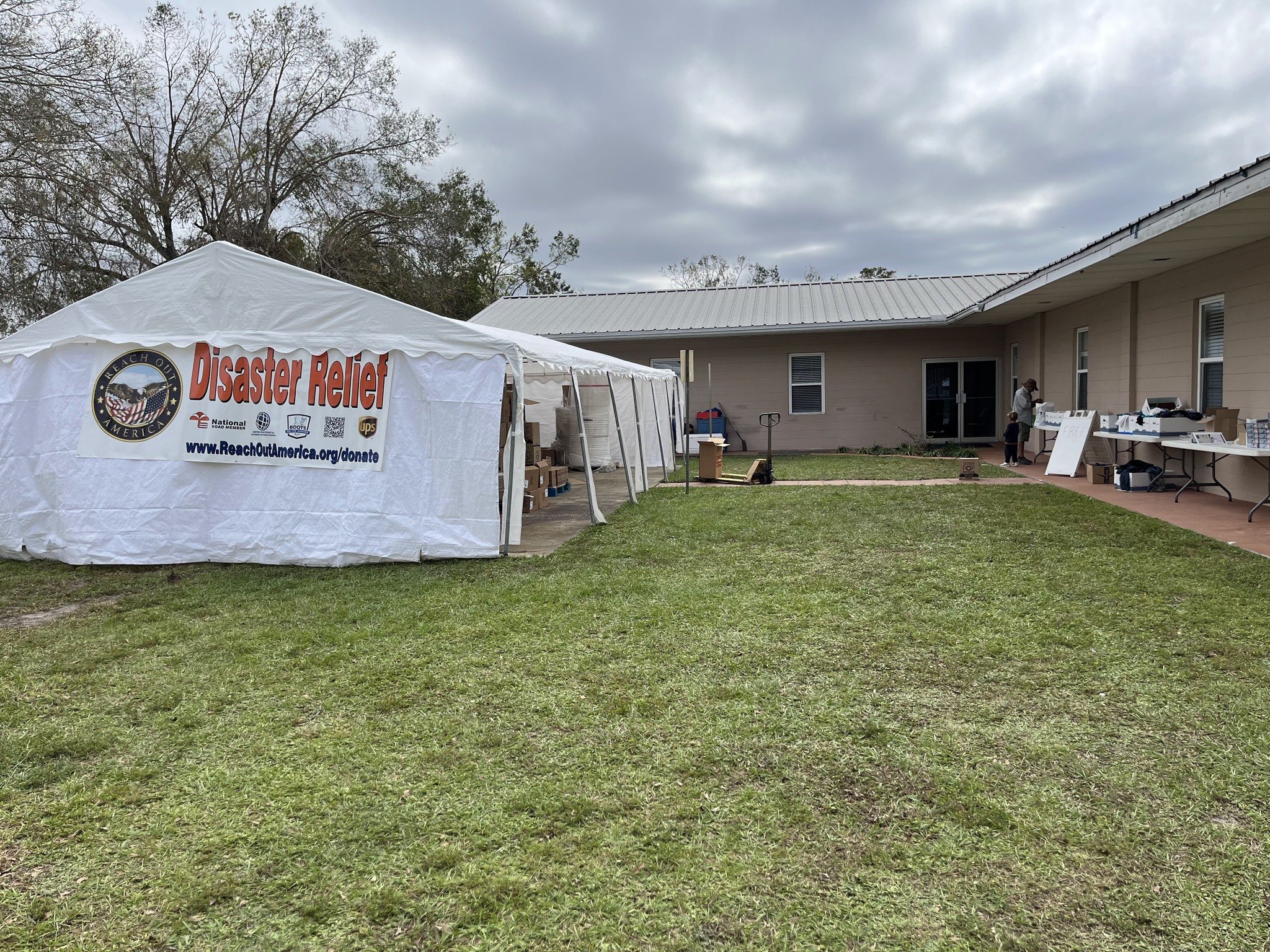 A large white tent with a sign that reads Disaster Relief and Reach Out America, with tables and supplies underneath, set up outside a beige building on a grassy area, with cloudy skies overhead.